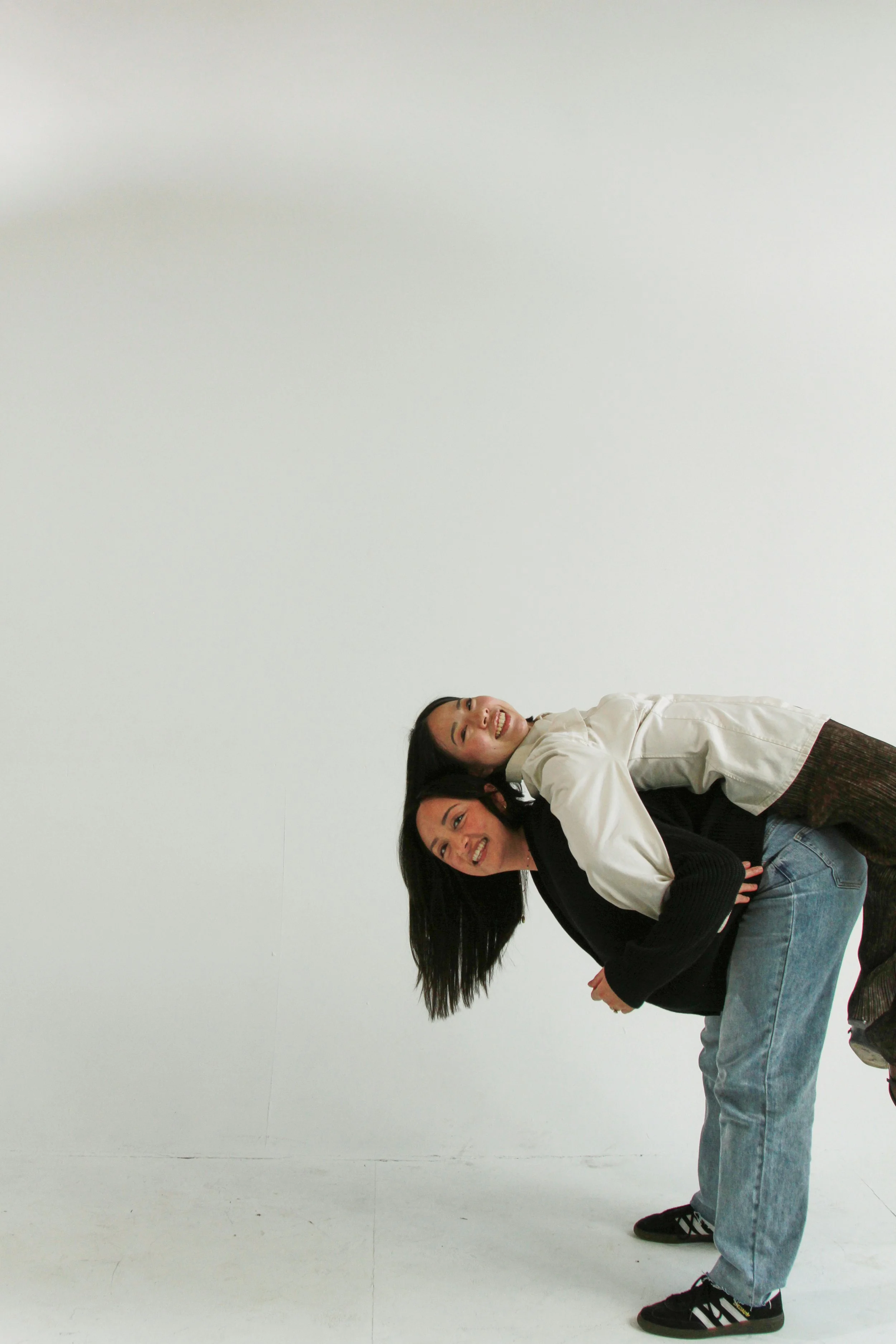 Two women are smiling and leaning over each other, standing in a minimalistic white room.