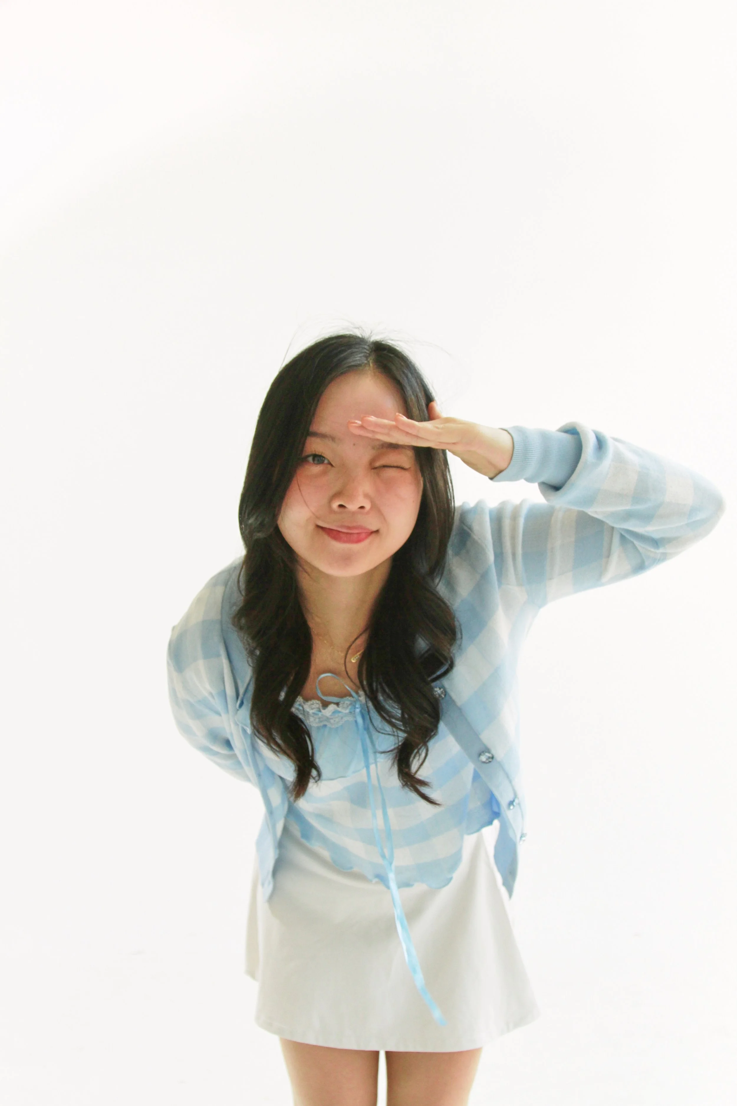 Young woman with long dark hair dressed in a pastel plaid button-up shirt and light-colored skirt, saluting and smiling against a plain white background.