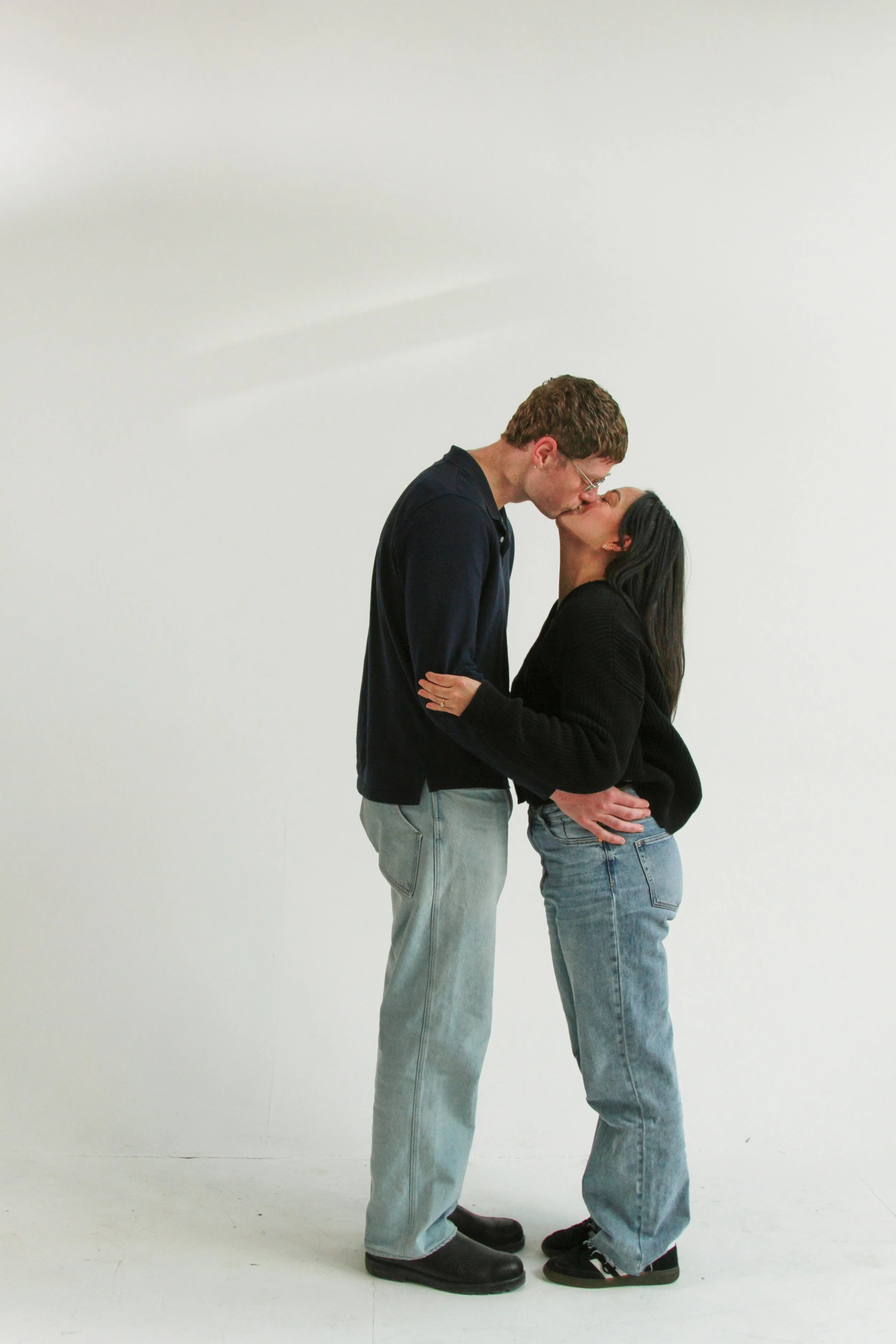 A young man and woman are standing close together, kissing, with their faces touching. The man is taller, wearing glasses, a black jacket, and light jeans. The woman has long dark hair, wearing a black sweater and slightly baggy jeans. They are stand