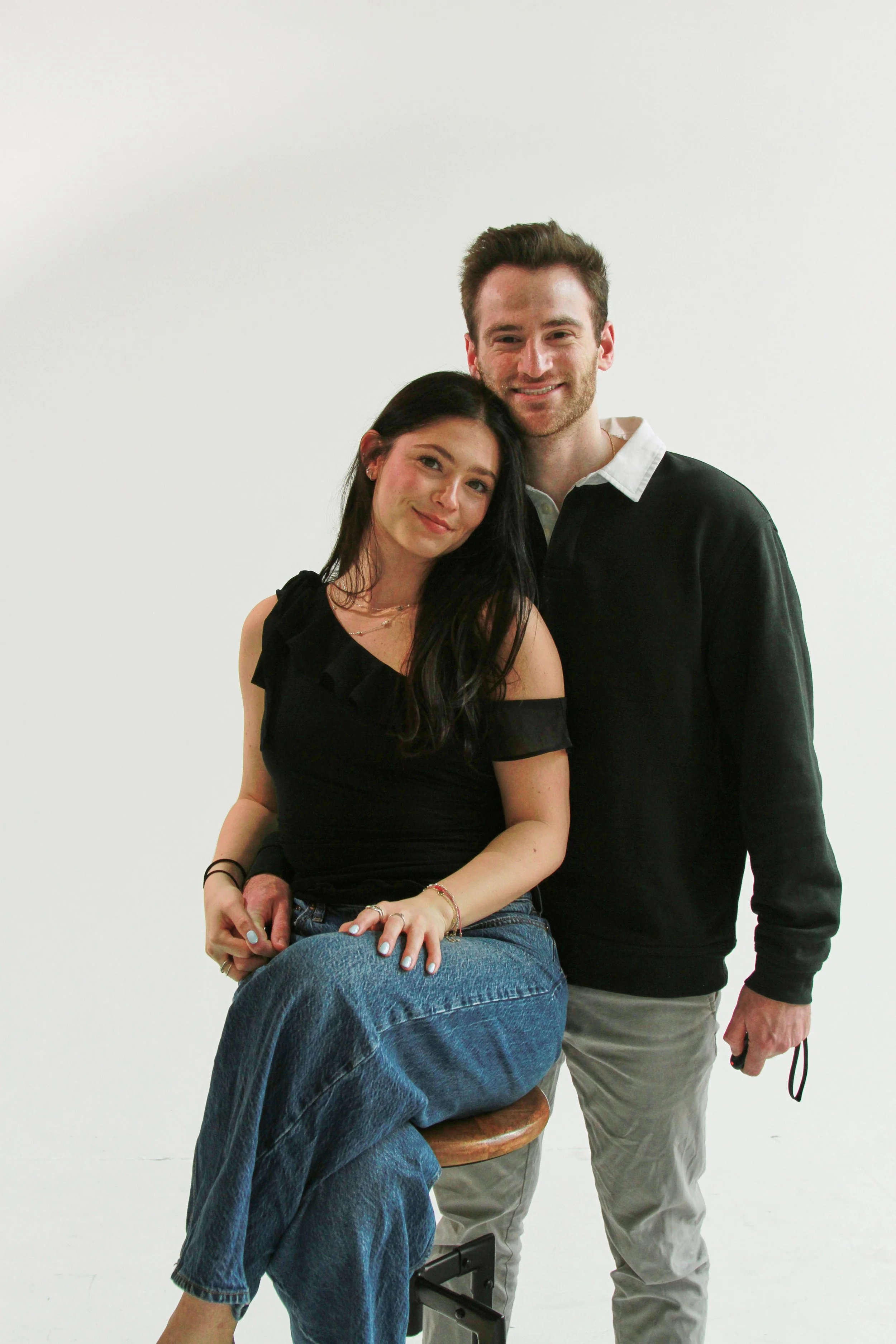 A young woman with long dark hair sitting on a stool, wearing a black top and blue jeans, with an older man standing beside her, both smiling.