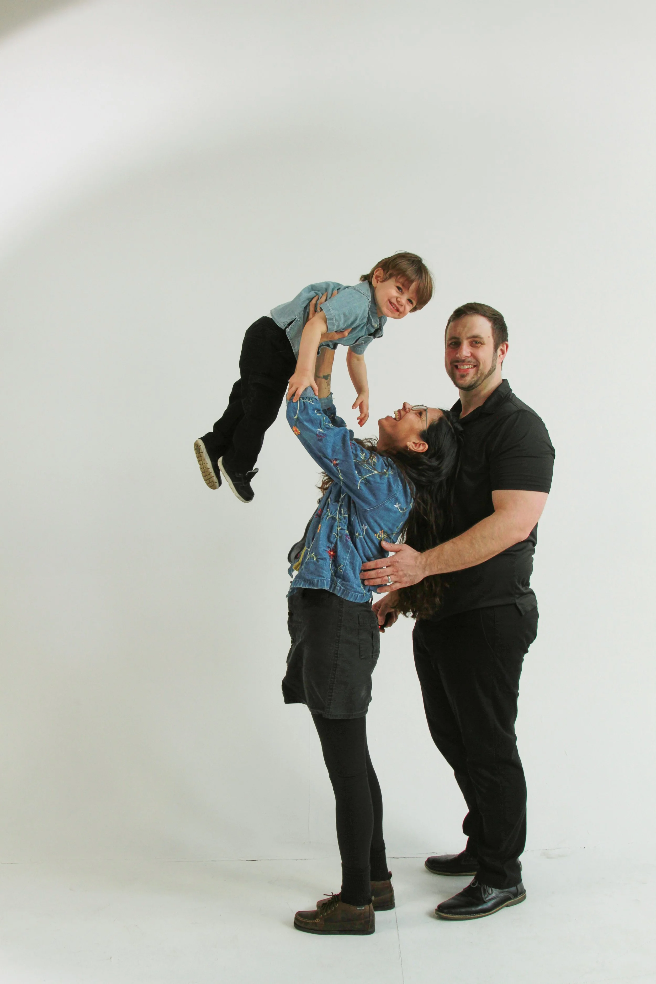 A woman lifts a young boy while a man supports the woman's waist, all smiling against a plain white background.