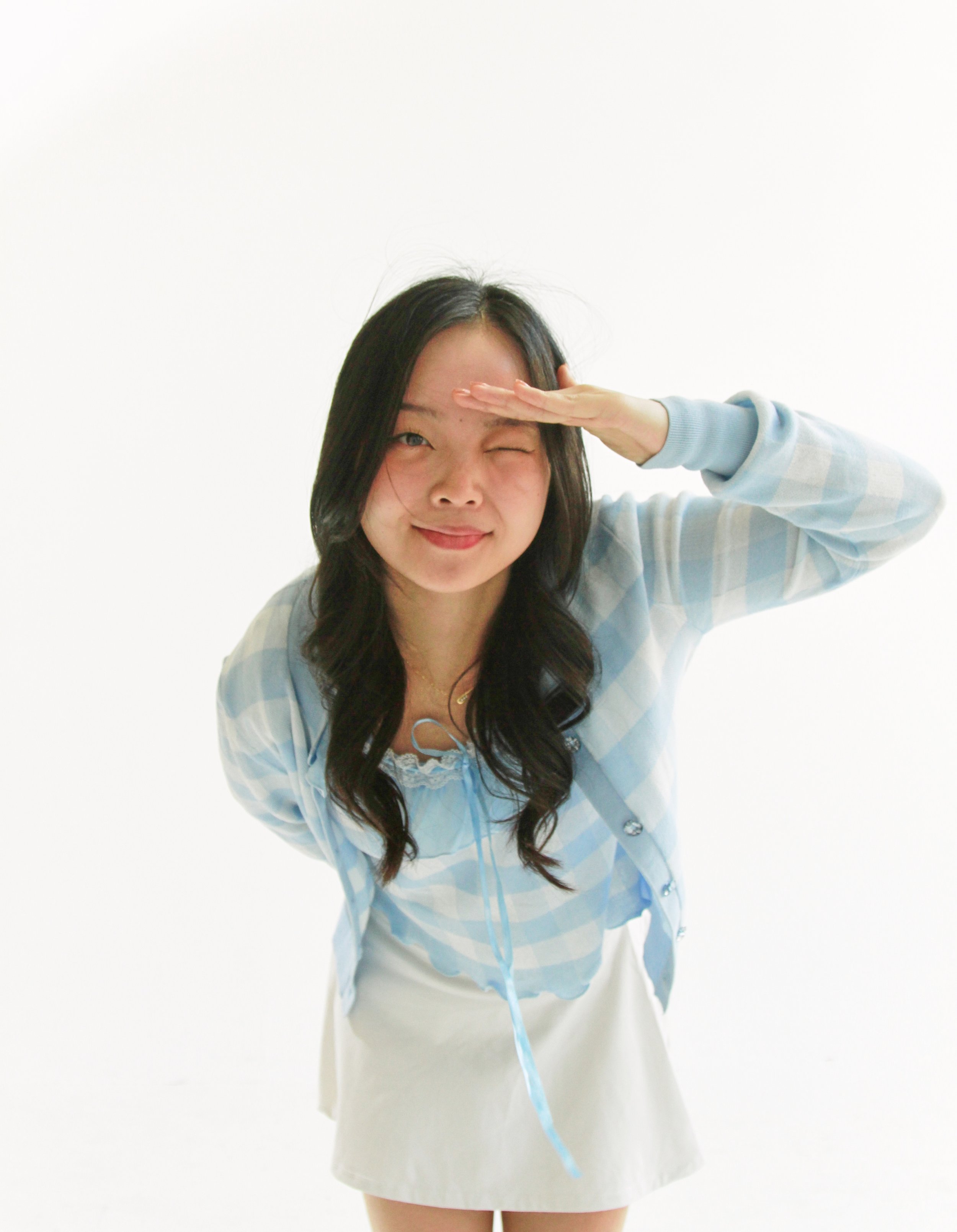 A woman with long dark hair, smiling and winking, salutes with her right hand while wearing a light blue plaid shirt and a white skirt, standing against a plain white background.