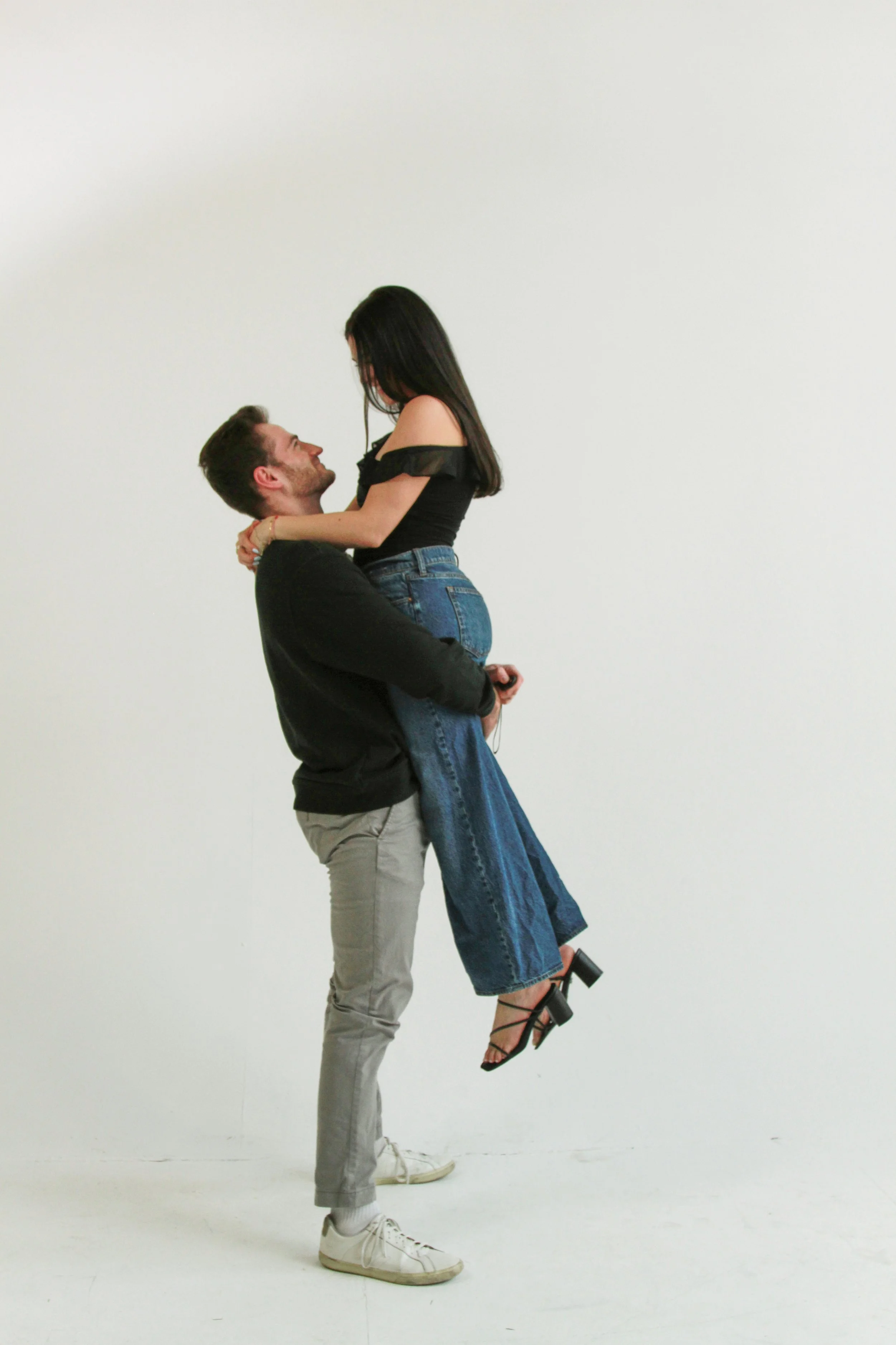 A man in gray pants and white sneakers holding a woman in a black top, denim skirt, and black high-heeled sandals in a dance pose against a plain white background.