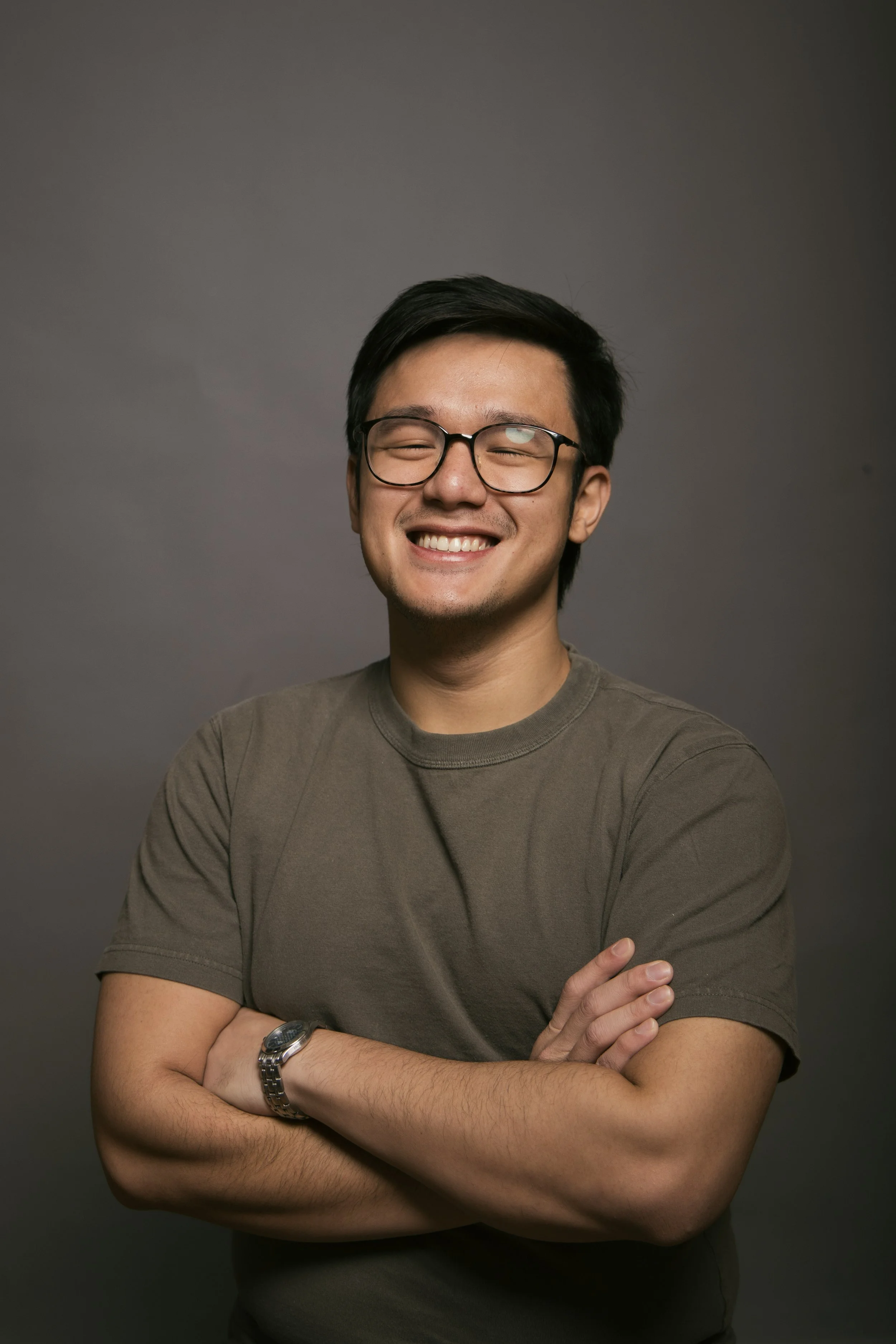 A young man with glasses smiling, arms crossed, wearing a watch and a brown t-shirt, posing against a dark grey background.