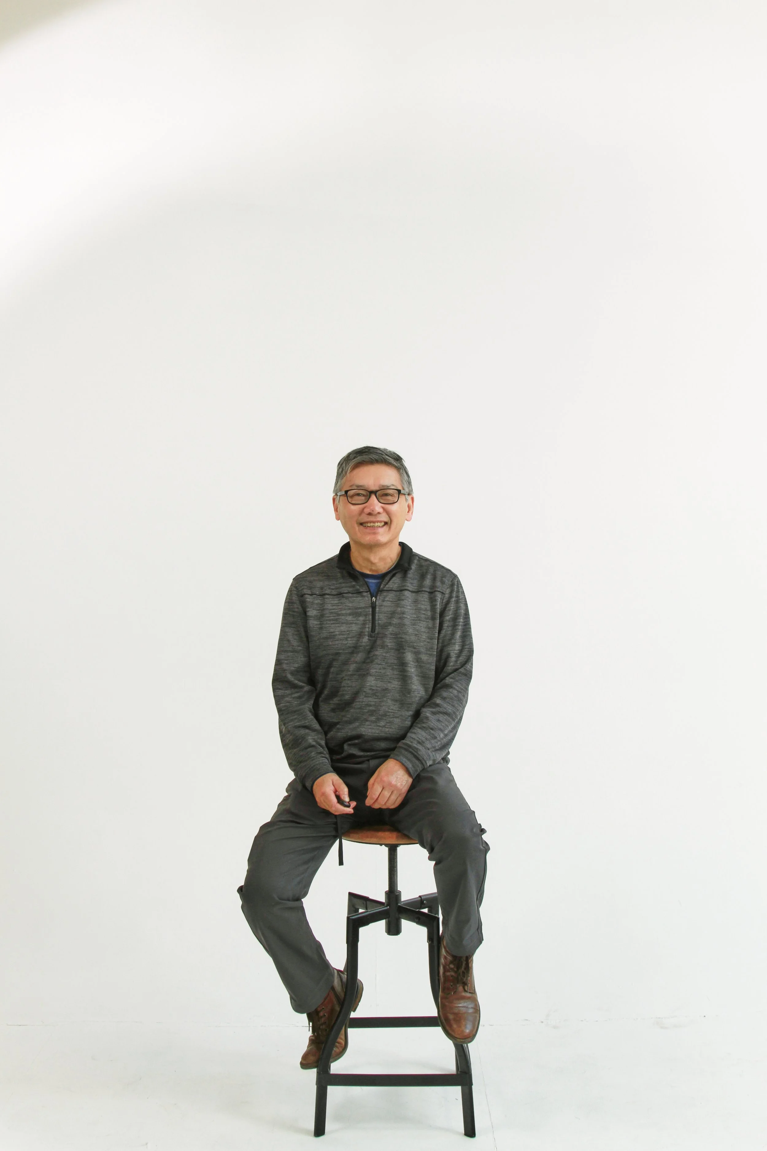 An older man with glasses smiling, sitting on a stool with one leg crossed, wearing a gray zip-up jacket and brown boots, against a plain white background.