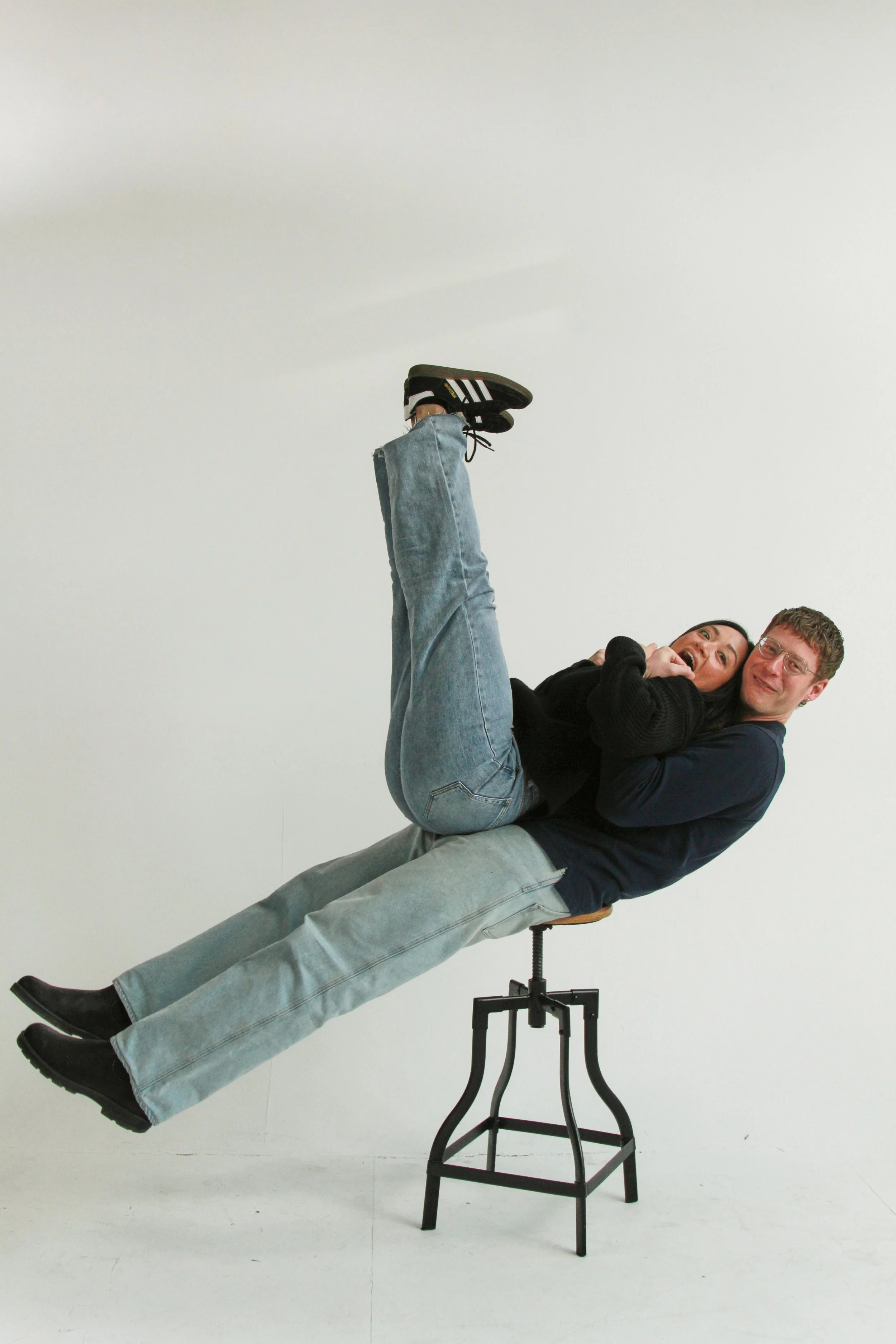 Two people, a woman and a man, lying on a chair with their legs raised, smiling, in a studio with a white background.