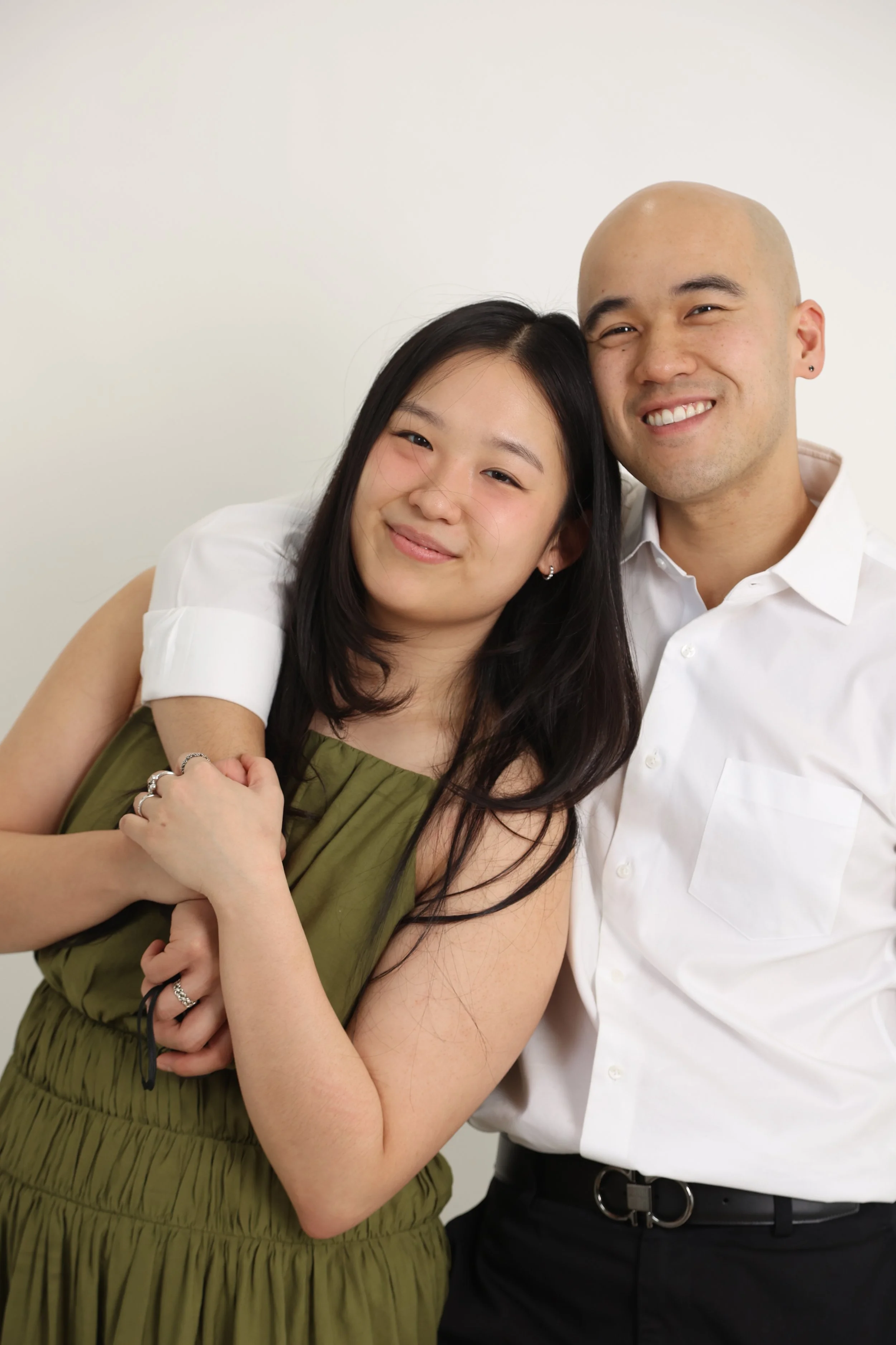 A young Asian woman with long dark hair is smiling and leaning against a bald young Caucasian man with a small earring, both are dressed casually and standing close together against a plain background.
