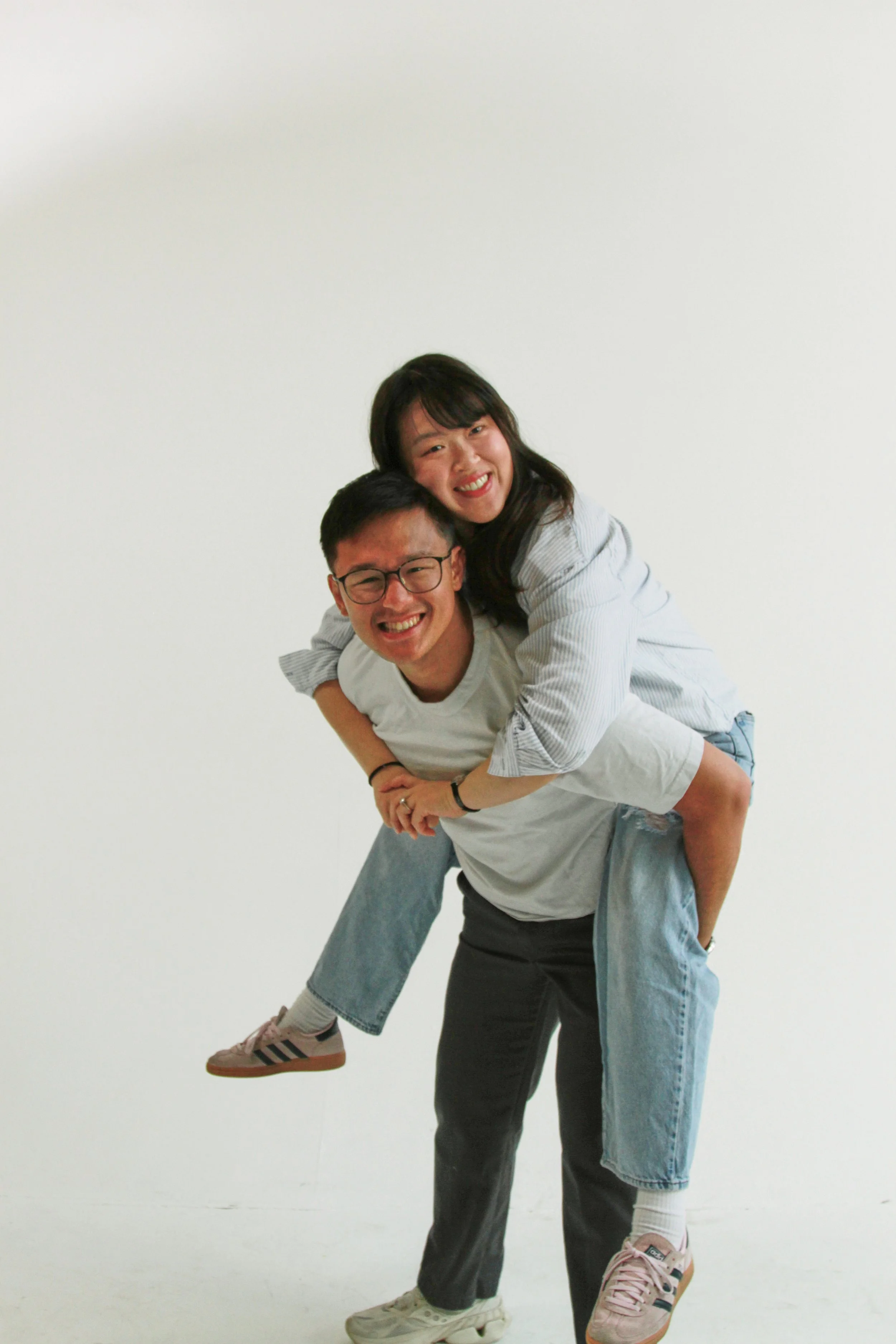 A young man with glasses and a woman with dark hair are playing piggyback against a plain white wall. The man is smiling widely, wearing a light gray t-shirt and black pants. The woman is smiling, leaning over the man's back with her arms around his 