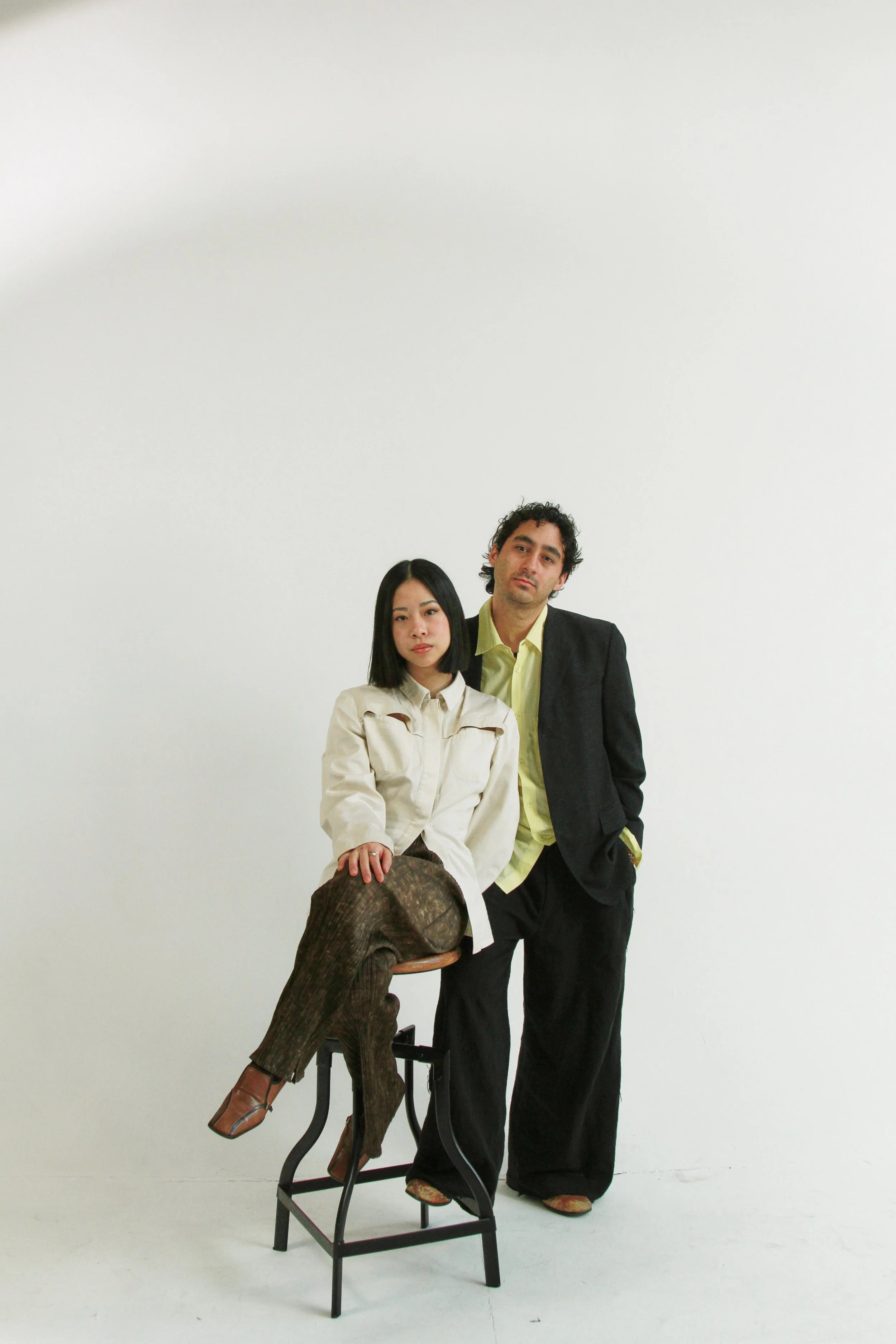 A man and a woman pose against a white wall. The woman is sitting on a chair with her left leg crossed over her right knee, wearing a cream-colored jacket and brown patterned pants. The man stands next to her, dressed in a black blazer and yellow shi