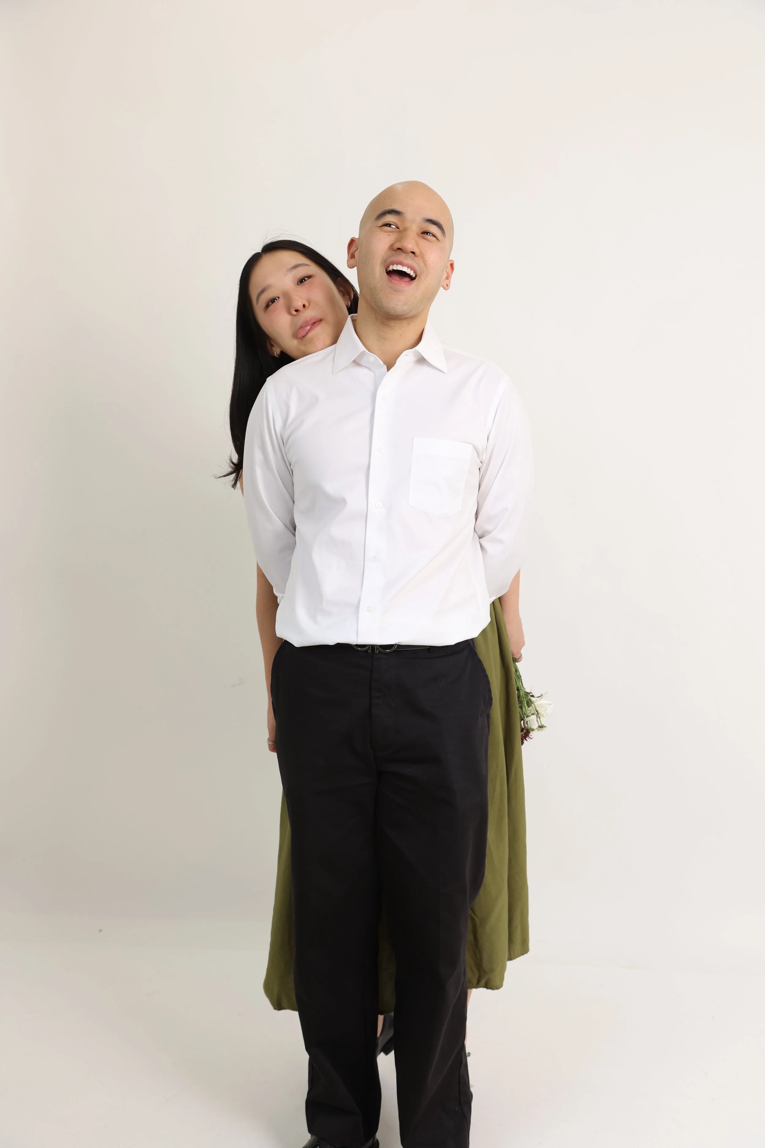 A man and a woman stand against a plain white wall, with the woman resting her head on the man's back and shoulders. The man is wearing a white dress shirt and black pants, and smiling as he looks upward and to the right. The woman is partially hidde