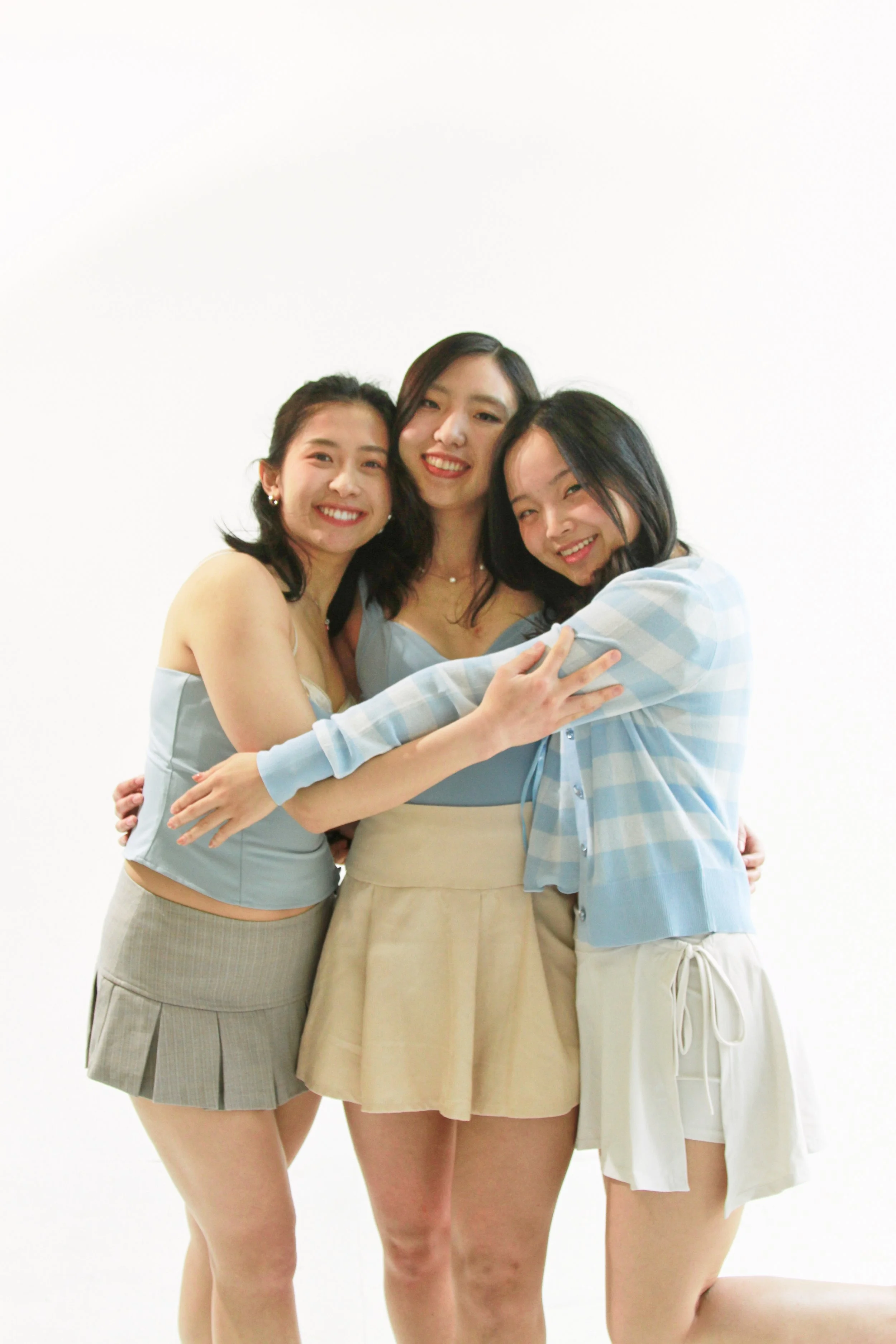 Three young women embracing and smiling at the camera, standing against a plain white background.