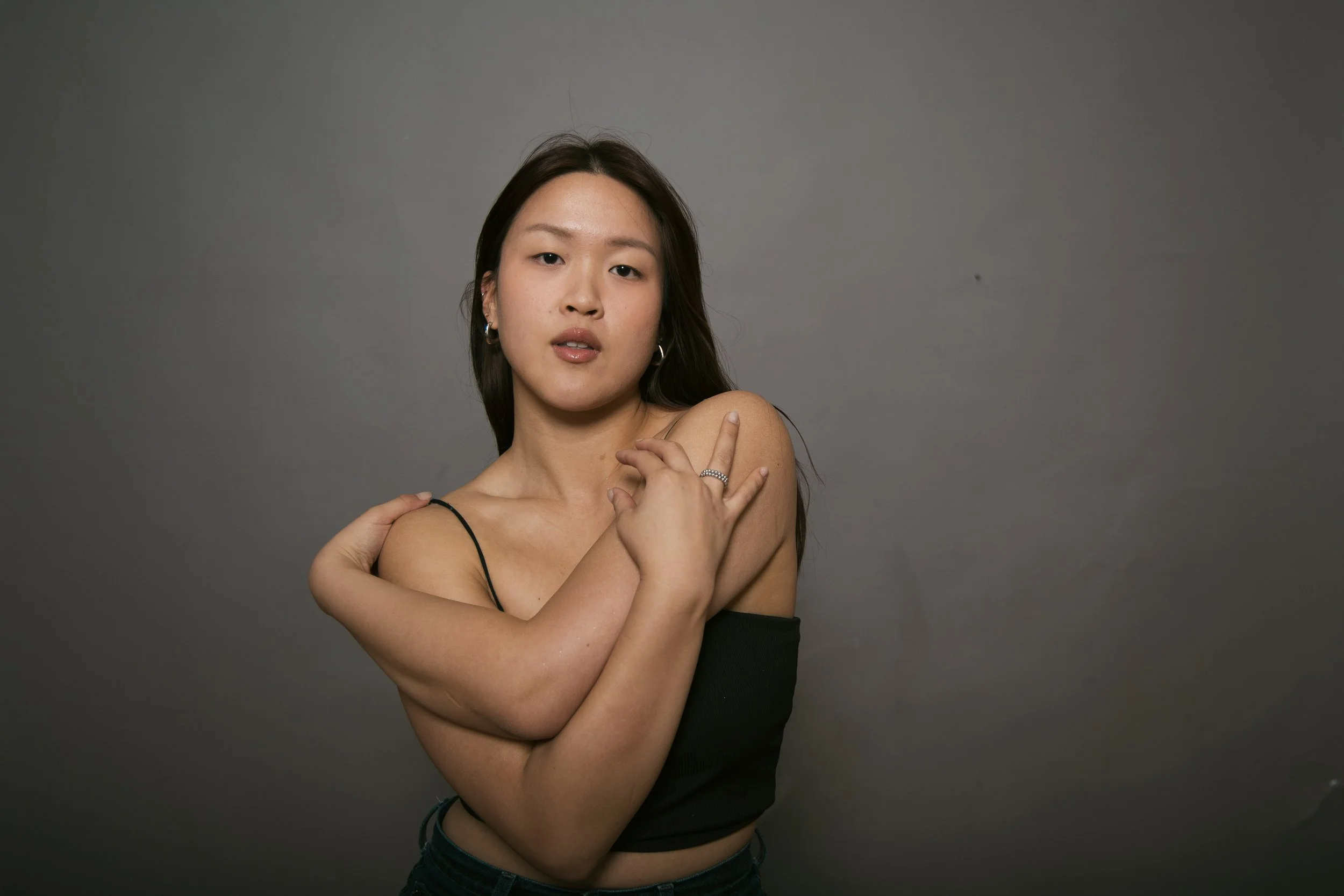A young woman with long dark hair, wearing hoop earrings, dressed in a black strapless top, standing against a plain gray background. She has her arms crossed, holding her shoulders with her hands, and gazes softly at the camera.