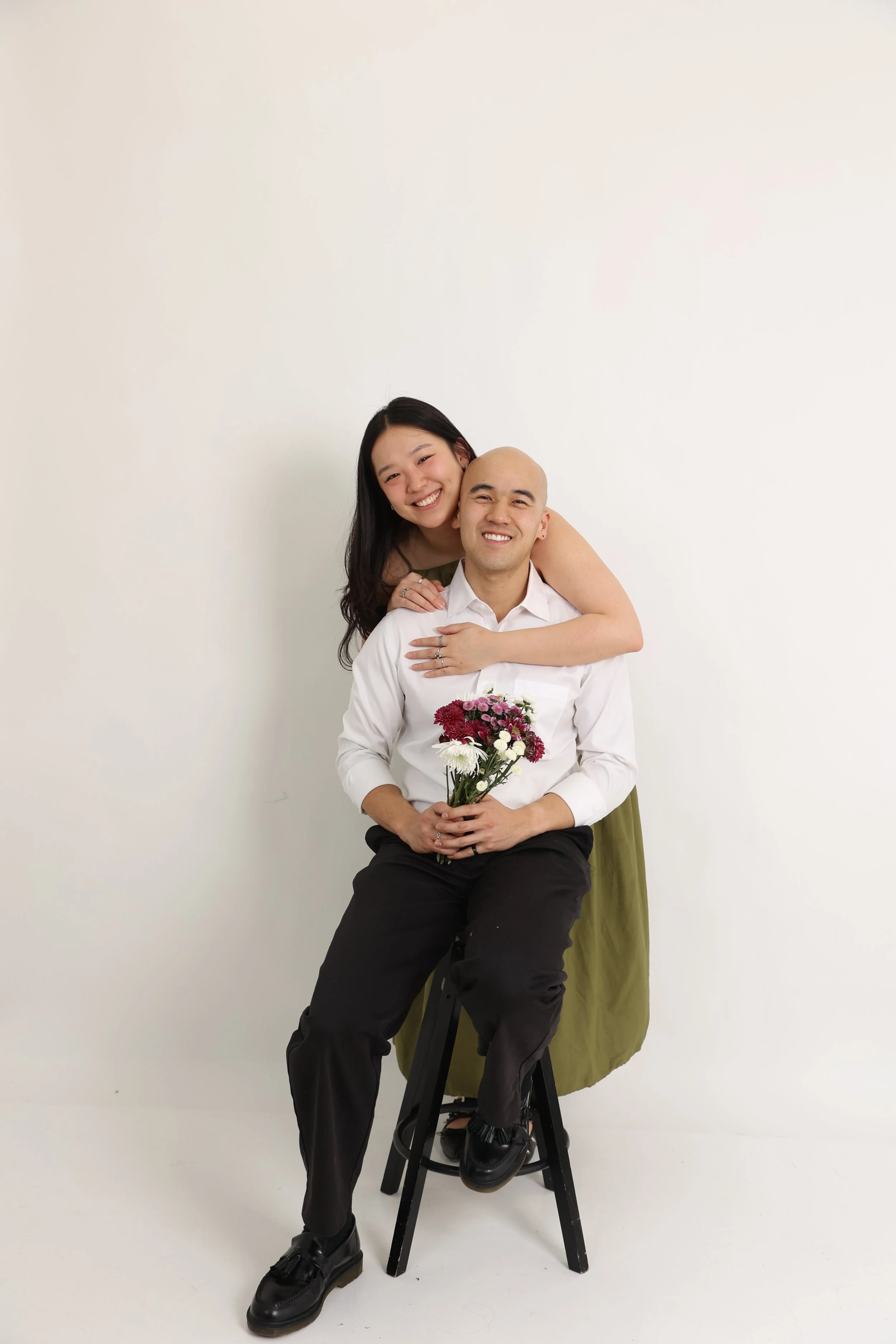 A smiling woman with long dark hair leaning on the shoulders of a seated man with a shaved head, both smiling. The woman has her arms around the man's shoulders, and the man holds a bouquet of flowers, wearing a white shirt. They are positioned again