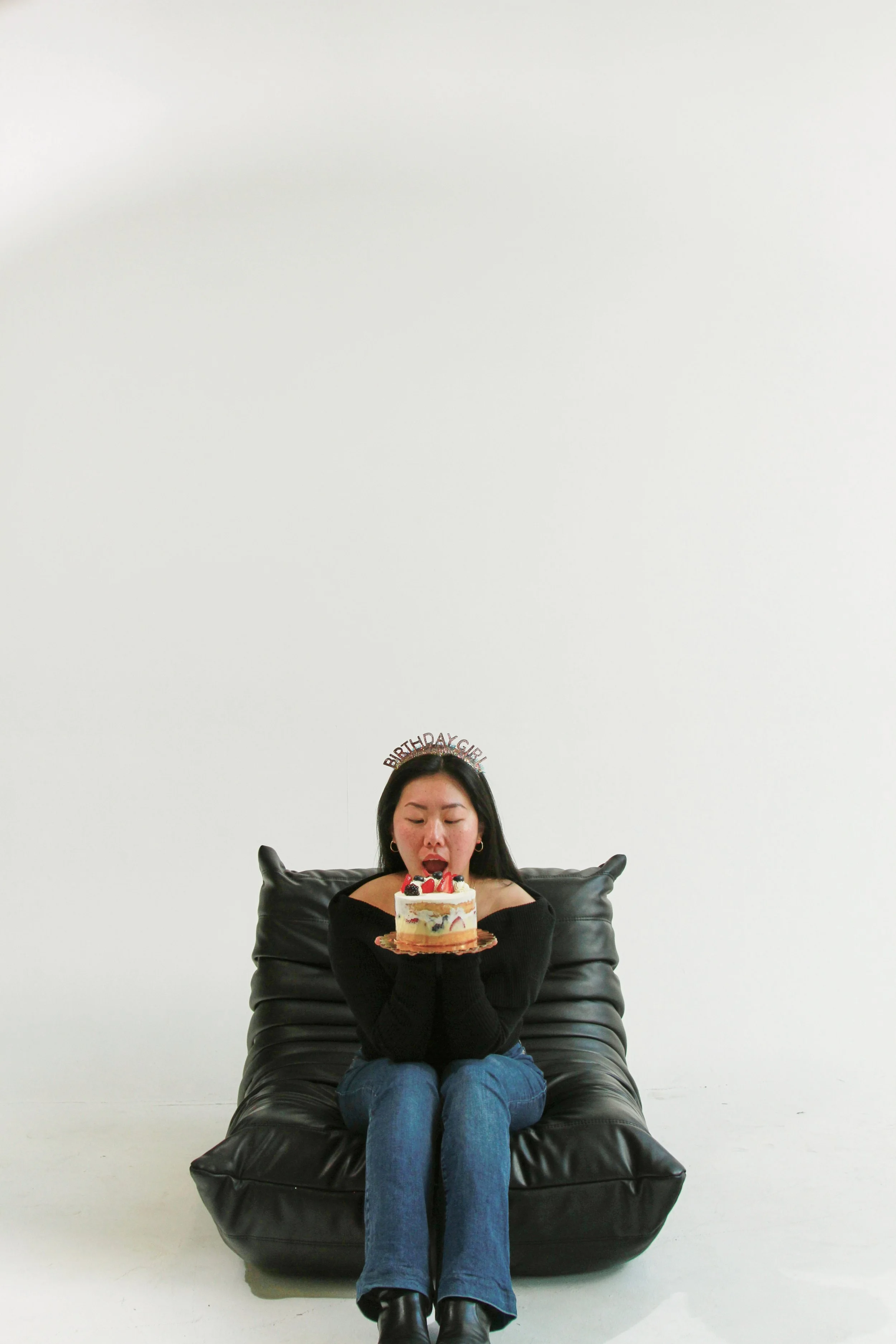 A woman sitting on a black leather chair, holding a birthday cake with strawberries and cherries, in front of a plain white wall background. She has dark hair, wears earrings, and a black long-sleeve top. A birthday hat with the words 'BIRTHDAY GIRL'
