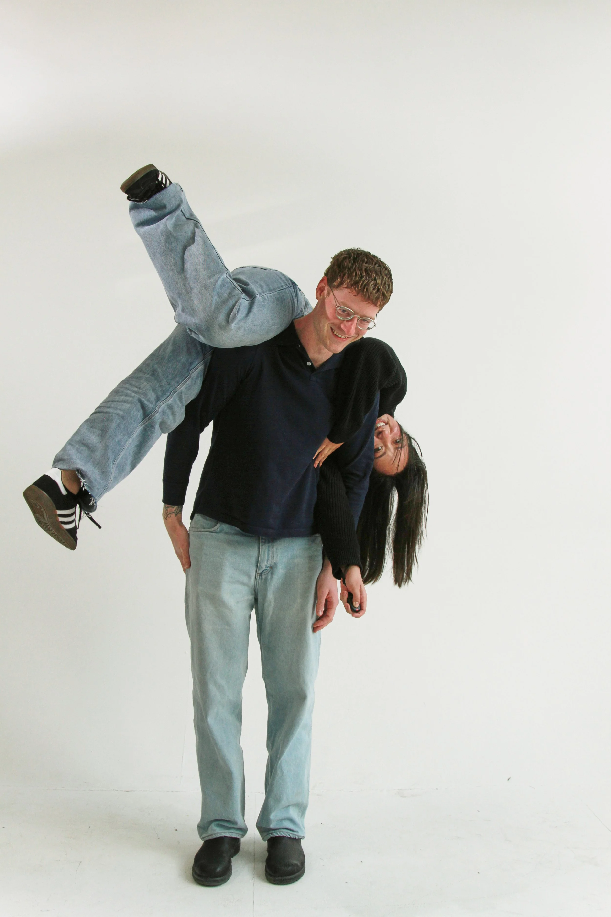 A man standing against a white background with a woman sitting on his shoulders, leaning forward with long black hair and glasses, smiling, both dressed casually in jeans and dark tops.