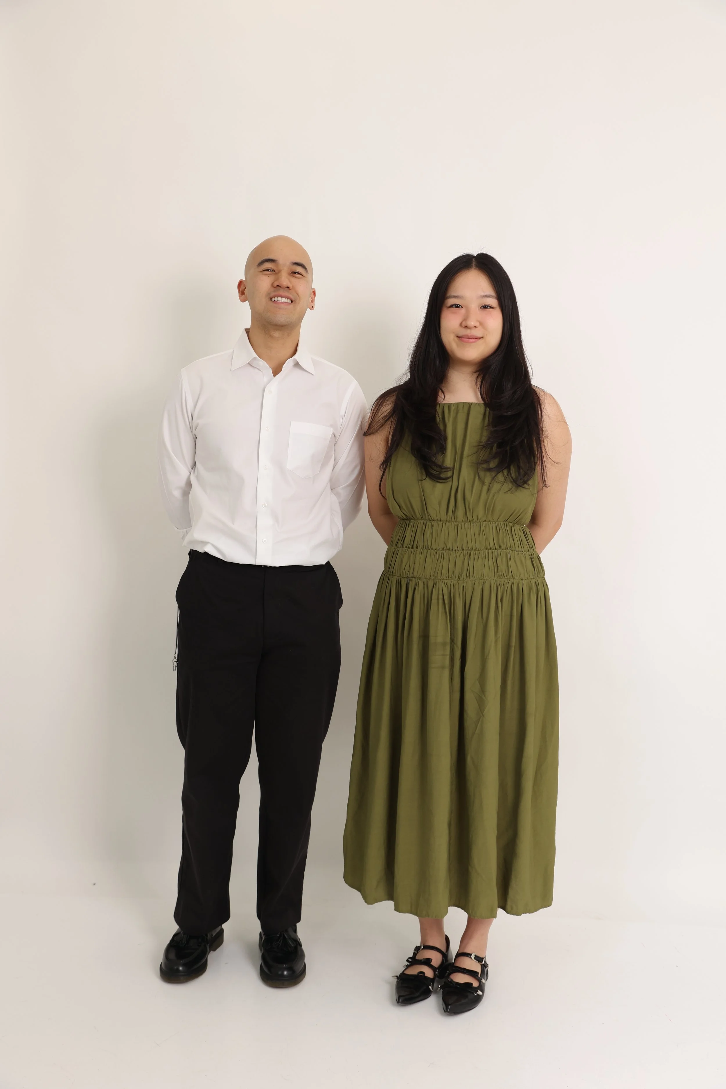 A man with a shaved head and a woman with long black hair standing against a plain white wall. The man is wearing a white dress shirt and black pants, while the woman is dressed in an olive green sleeveless dress with a gathered waist. They are both 
