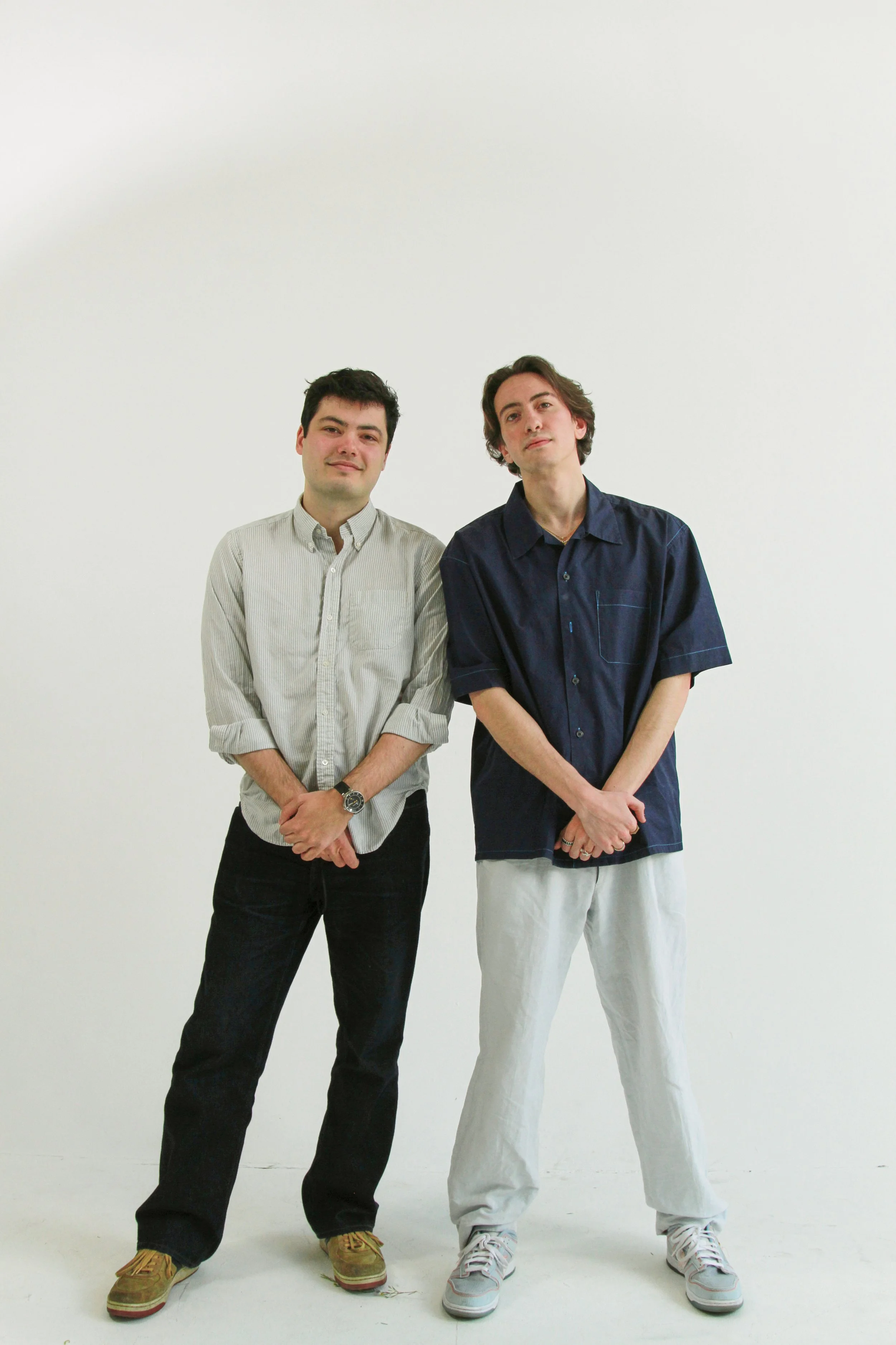 Two young men standing against a white background, dressed casually in button-up shirts and pants.
