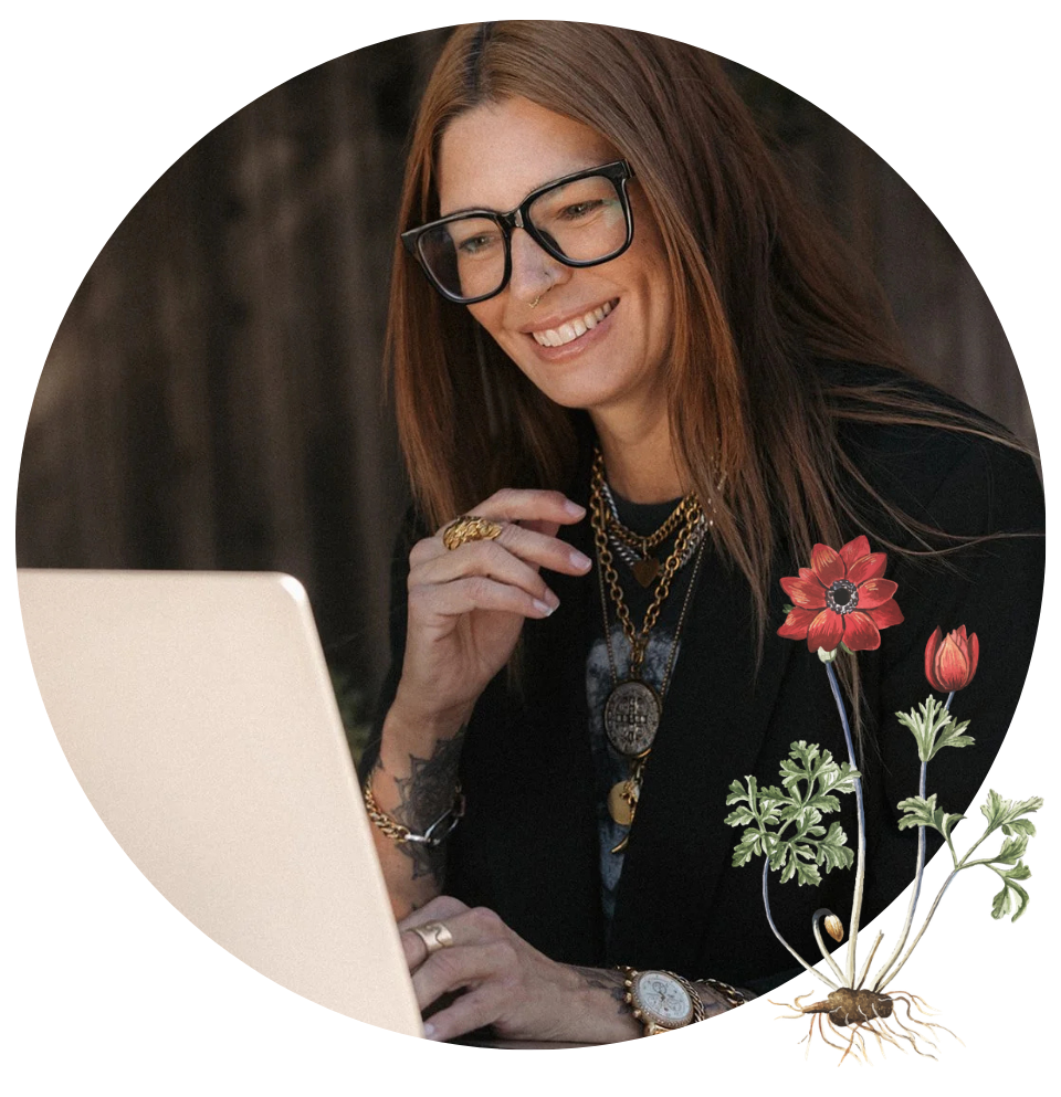 A woman with long red hair and glasses smiling while using a laptop, wearing layered necklaces and rings, with a dark wooden background and illustrated flowers and plants in the foreground.