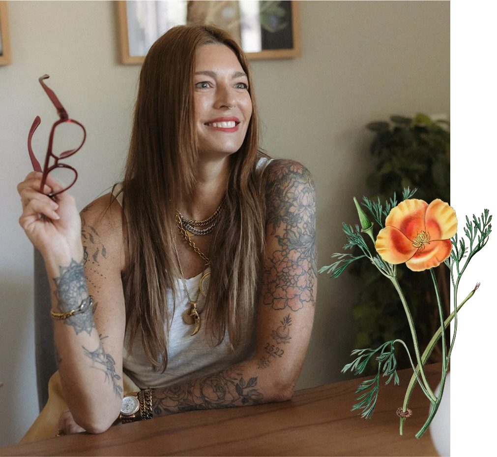 A woman with long reddish-brown hair, tattoos on her arms, and jewelry, sitting at a wooden table, holding a pair of glasses, smiling and looking to her right, with a blurred indoor background and an edited flower illustration on the right side of the image.