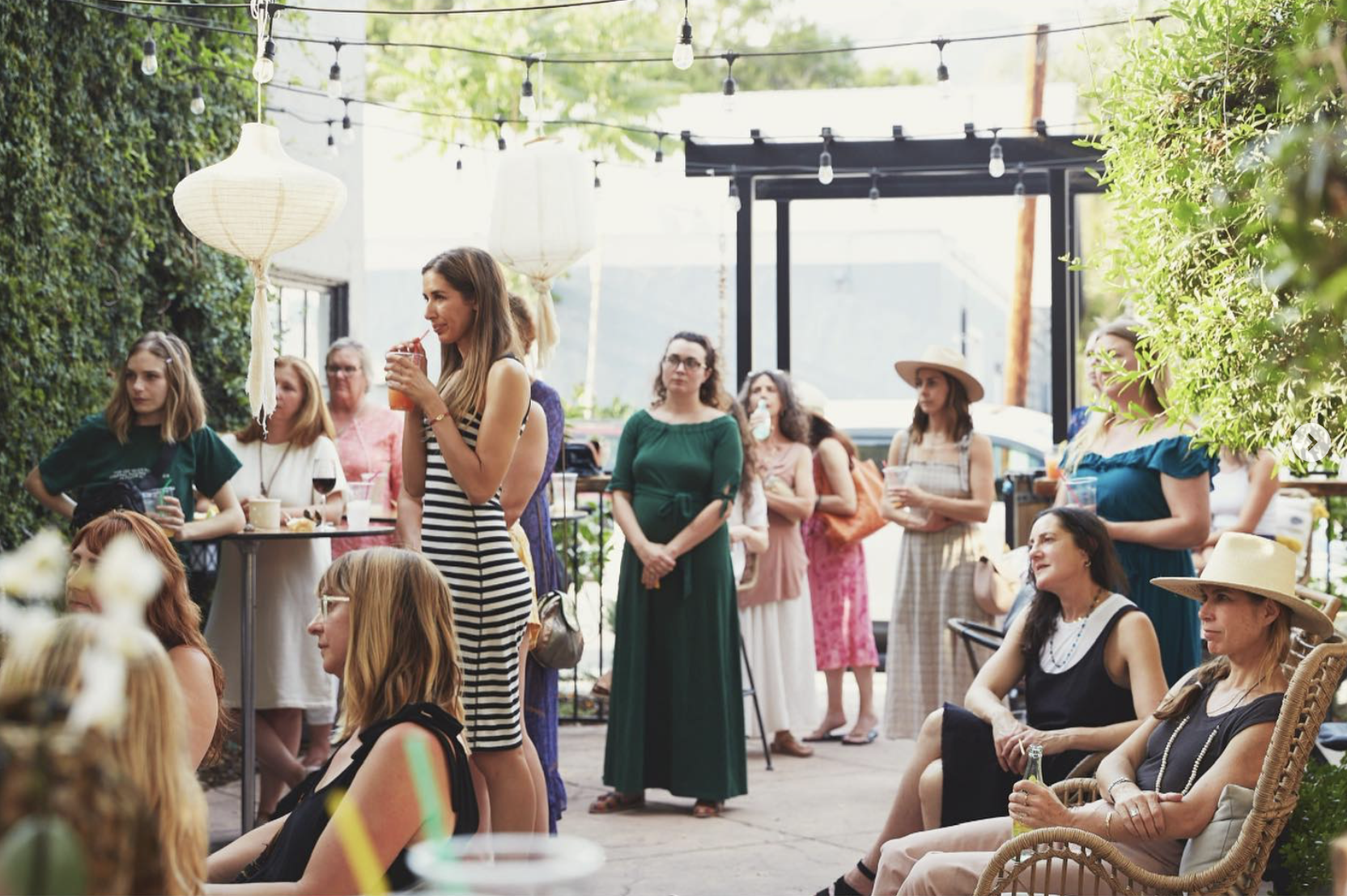 Women gathered at an outdoor social event on a patio with hanging lanterns and string lights, some sitting and others standing, engaged in conversation.