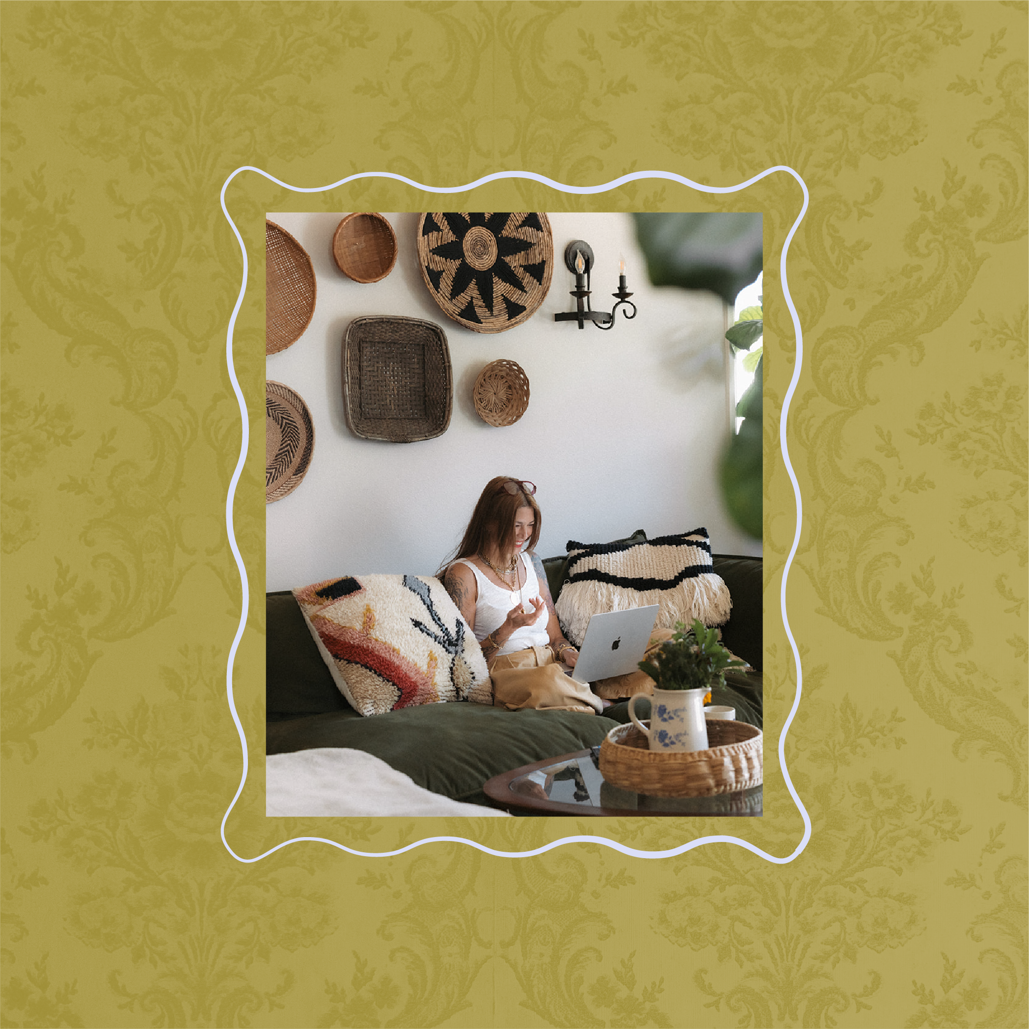 A woman sitting on a sofa with colorful pillows, using a laptop, with a coffee table in front holding a pitcher and cups. The background wall displays various woven baskets and a wall sconce.
