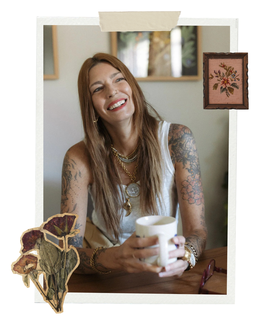 A smiling woman with tattoos, long light brown hair, wearing jewelry, sitting at a table, holding a white mug.