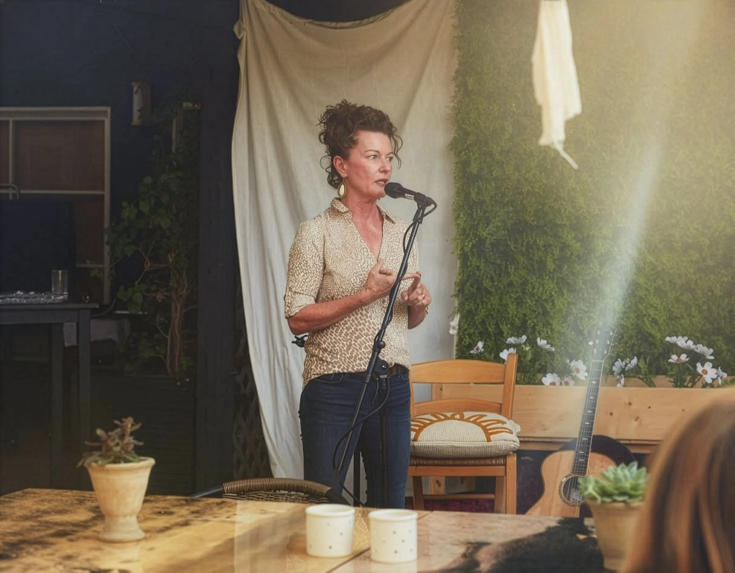 A woman singing or speaking into a microphone in an indoor setting with plants and a guitar nearby.