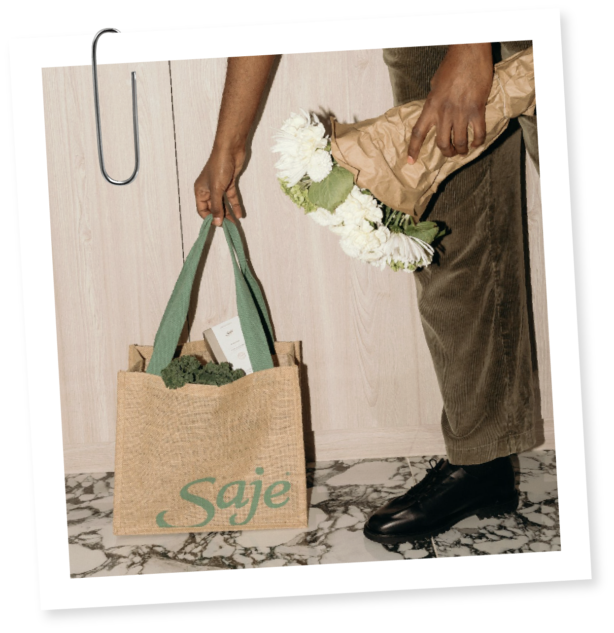 Person holding a paper bag with vegetables and a bouquet of white flowers, standing on marble floor in front of a light-colored wooden wall.