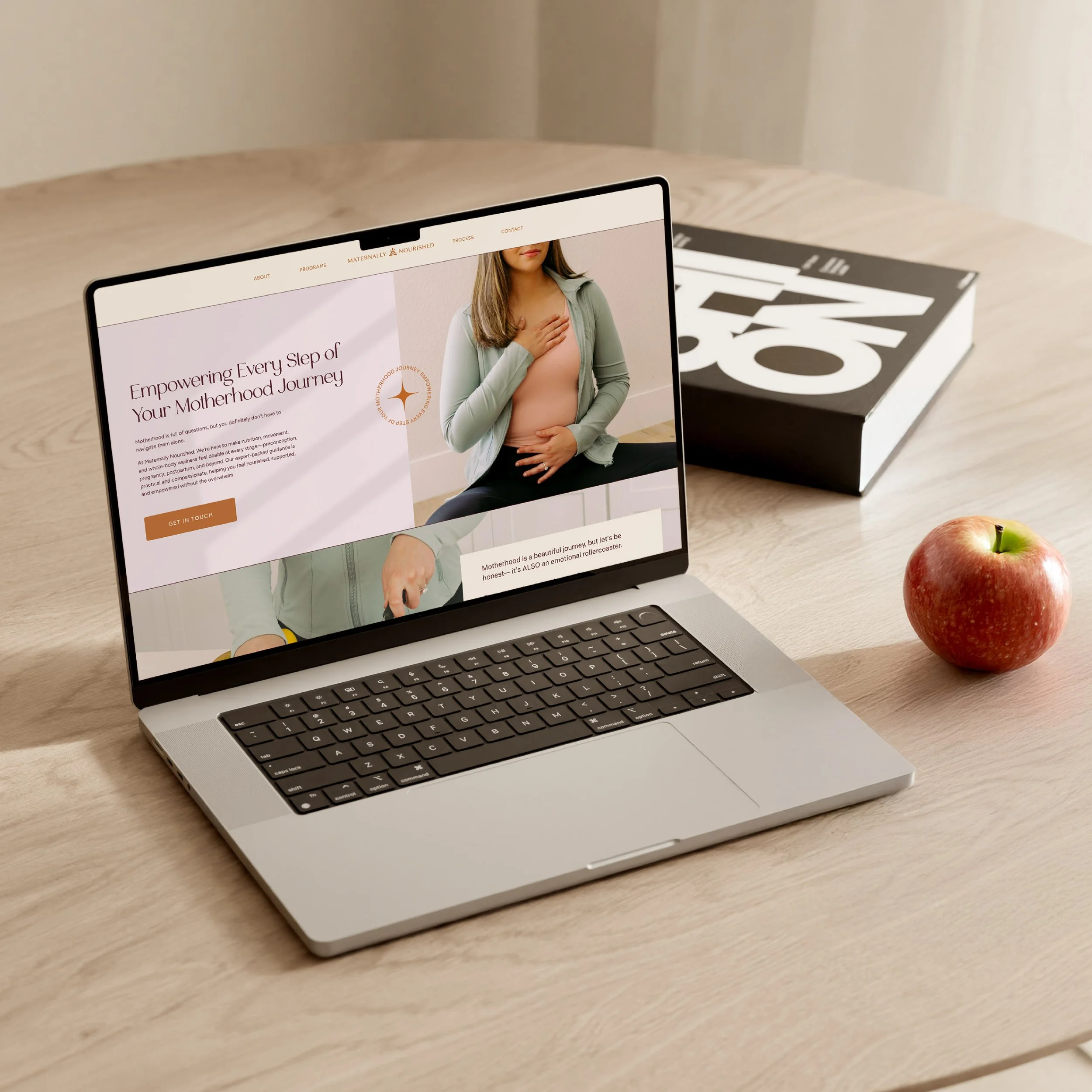 A laptop displaying a webpage about motherhood and pregnancy on a wooden table, with a magazine and apples nearby.