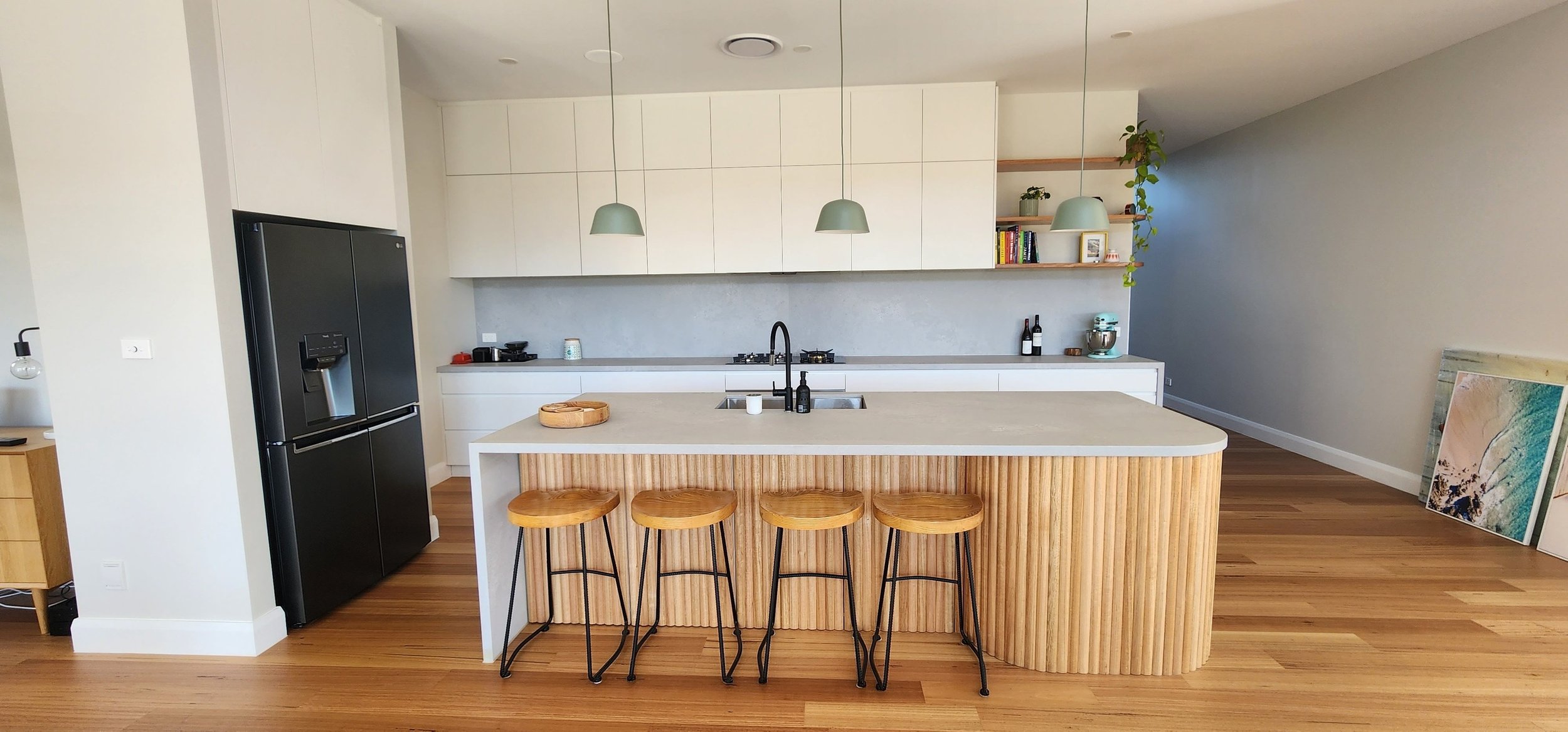 Modern kitchen with a white countertop island, four wooden bar stools with black metal legs, black sink faucet, white cabinetry, and open wooden shelves with books and decor, hardwood floors, and paintings leaning against the wall.