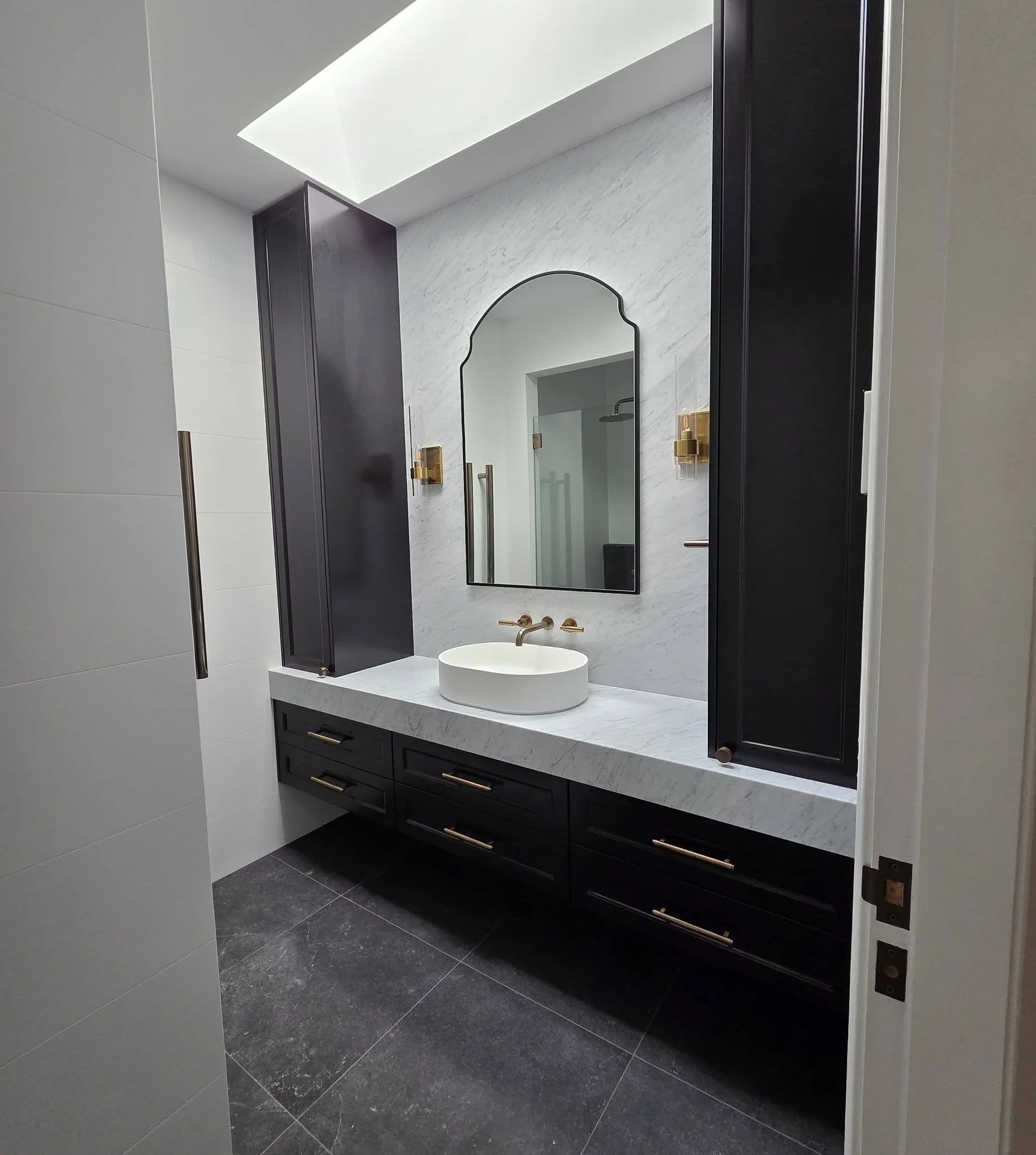 A modern bathroom vanity area featuring a white oval vessel sink, a gold faucet, a mirror with a dark frame, black cabinetry with gold handles, and wall-mounted gold fixtures, with a large white flooring tile and a marble wall.