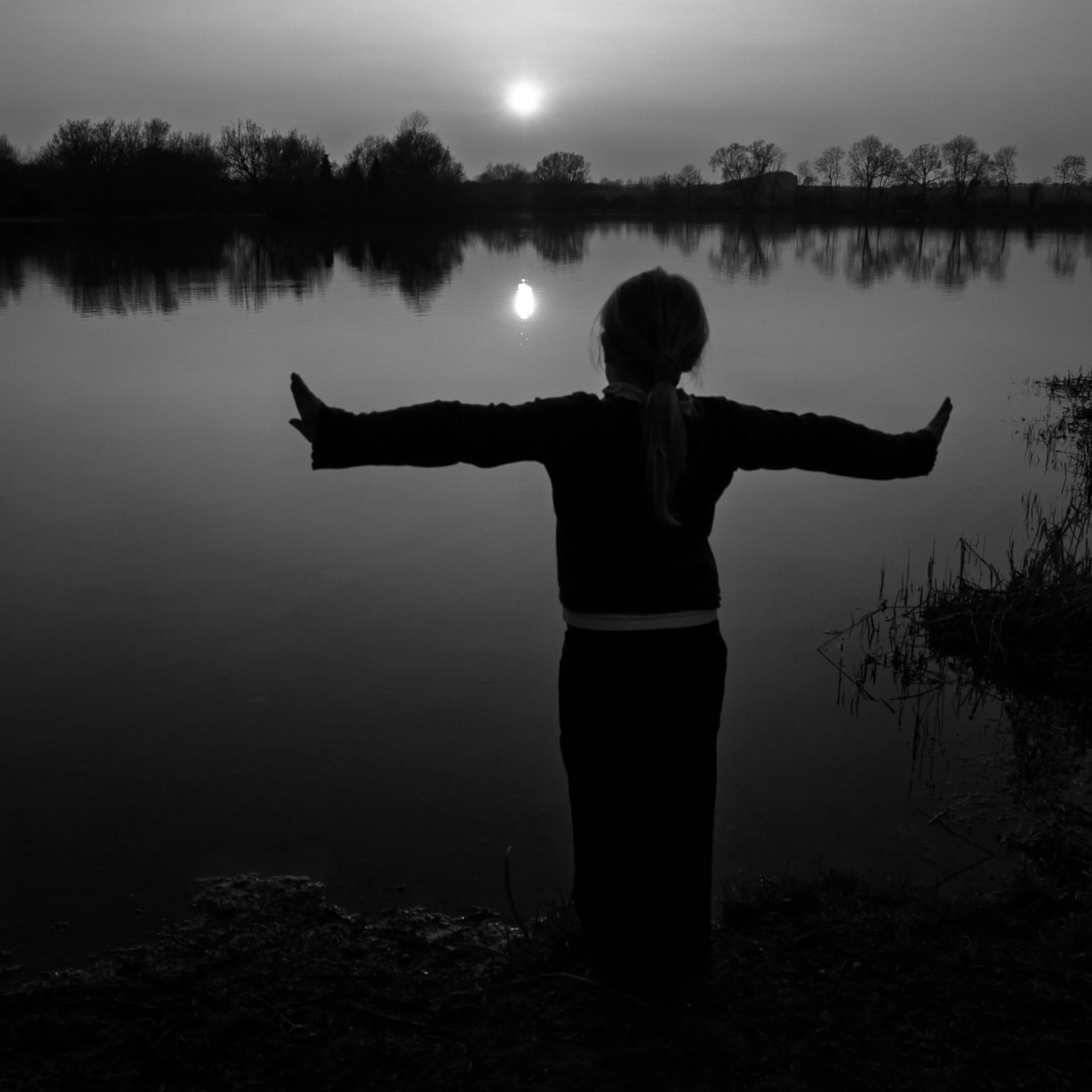 Black and white photo of a small girl standing by the edge of a lake at sunset, arms stretched out to either side.