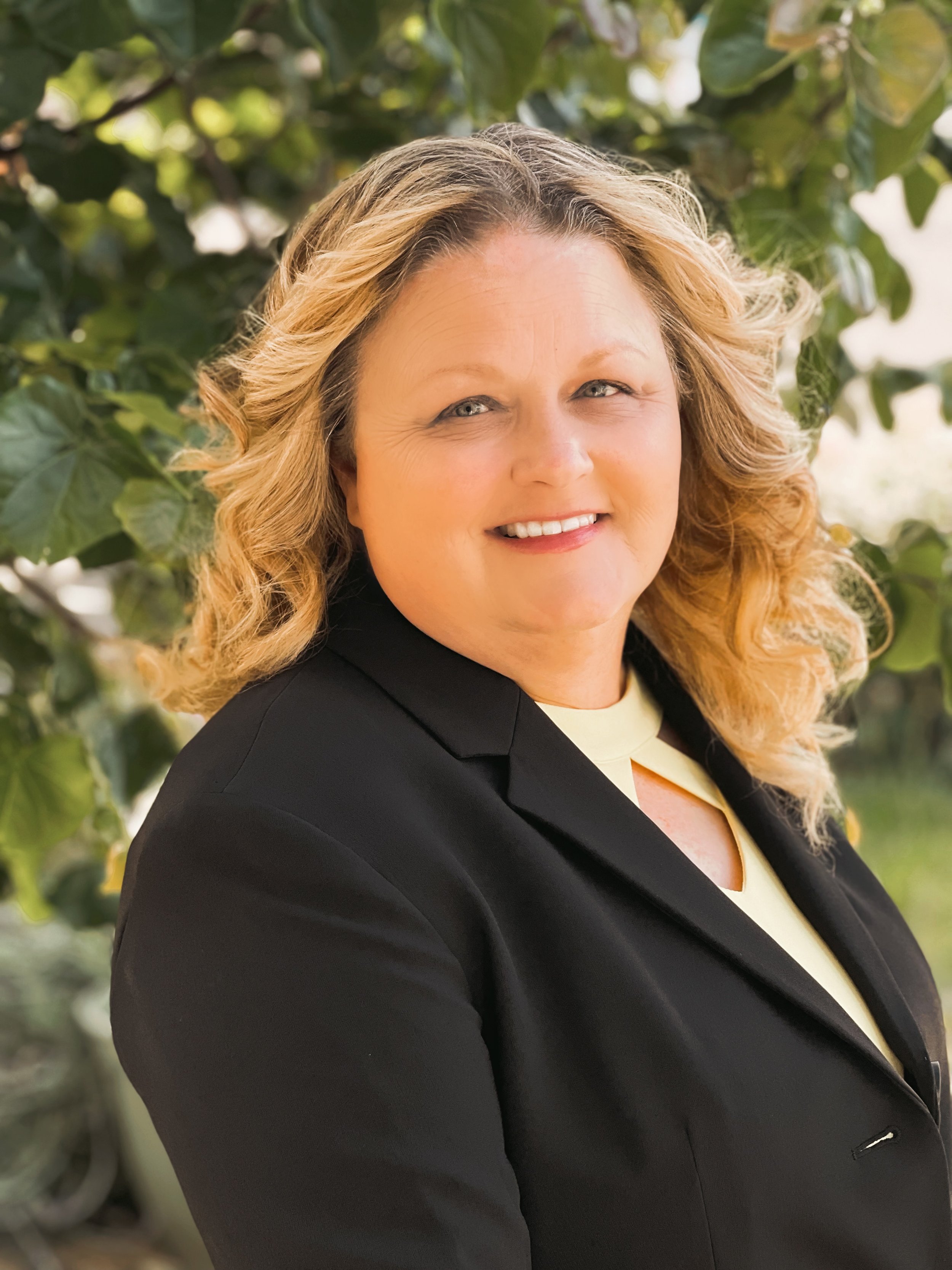 A woman with blonde, wavy hair wearing a black blazer and light yellow top, standing outdoors with green leaves in the background.