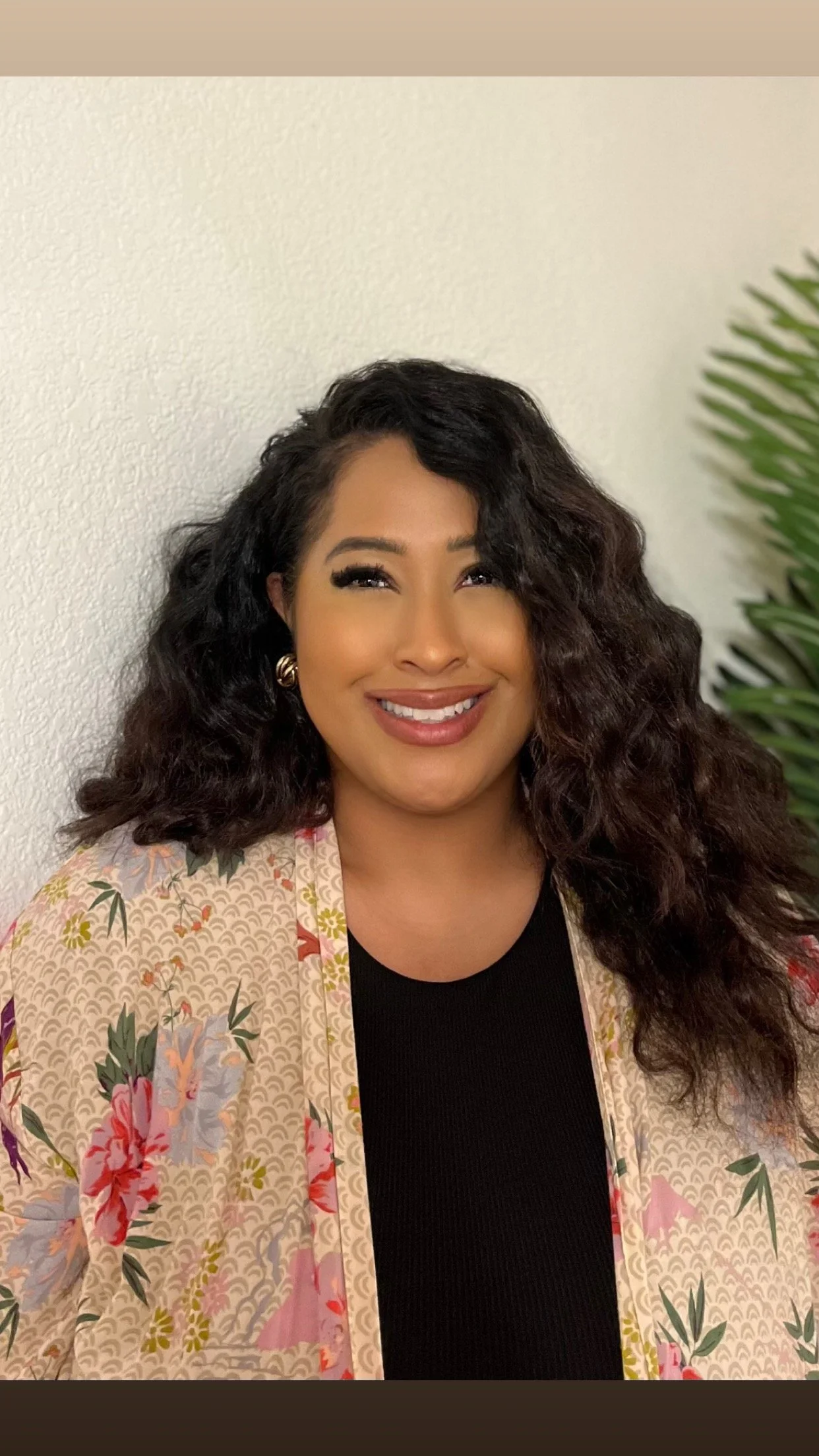 A woman with dark, curly hair smiling, wearing a black shirt and a floral patterned blazer, with a textured white wall and a green plant in the background.
