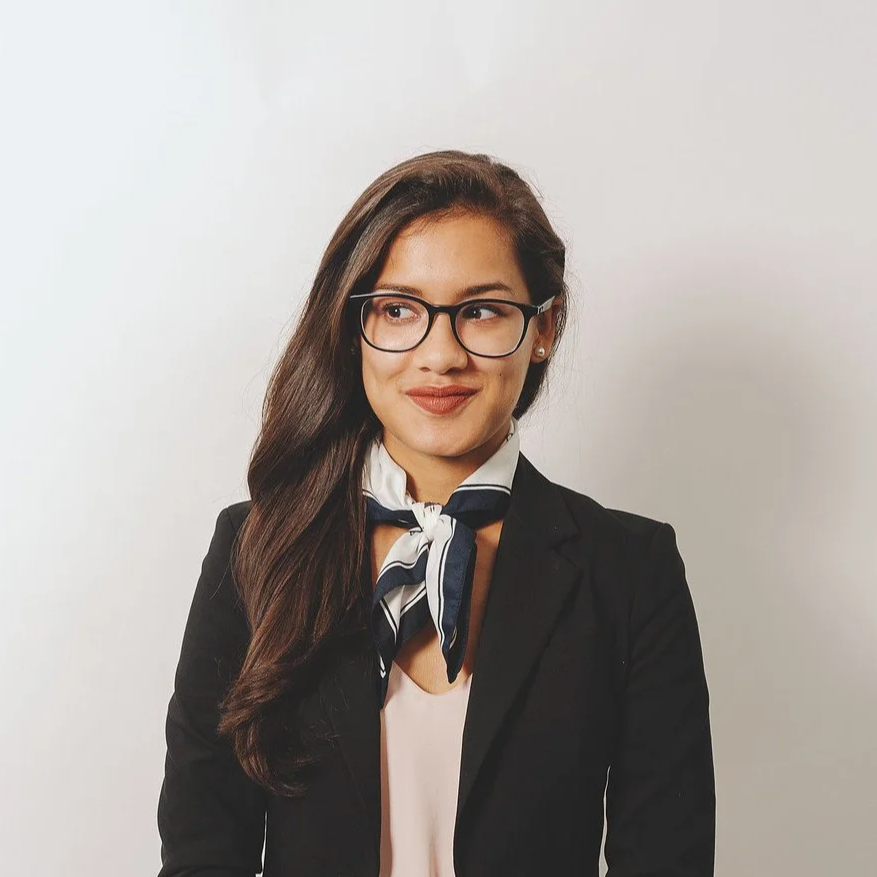 A portrait photo of a half-Filipina woman with long hair and glasses. She is wearing a black blazer and a white-and-blue silk necktie, and she is staring to her right.