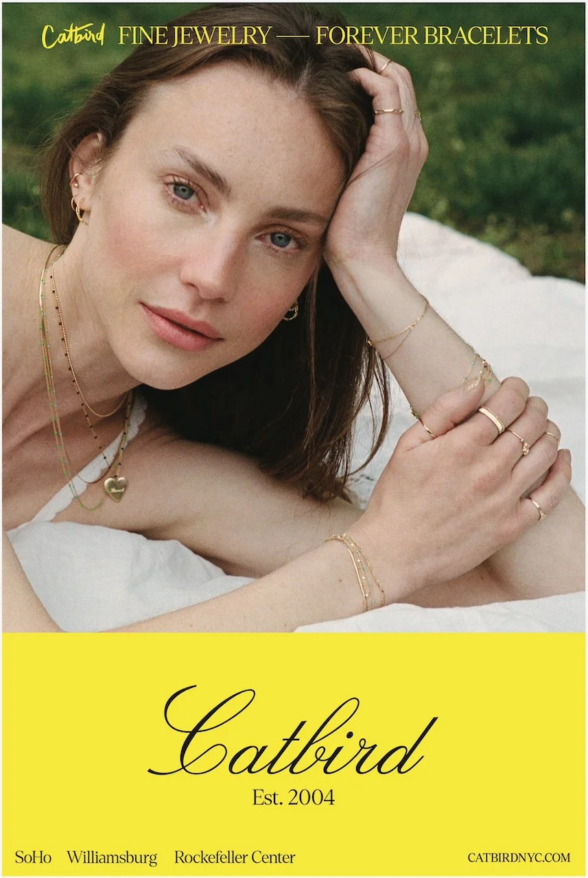 A woman with blue eyes and brown hair lying on a white surface outdoors, wearing multiple gold jewelry pieces including rings, necklaces, earrings, and bracelets. She is resting her head on her hand and looking at the camera. The background is green 