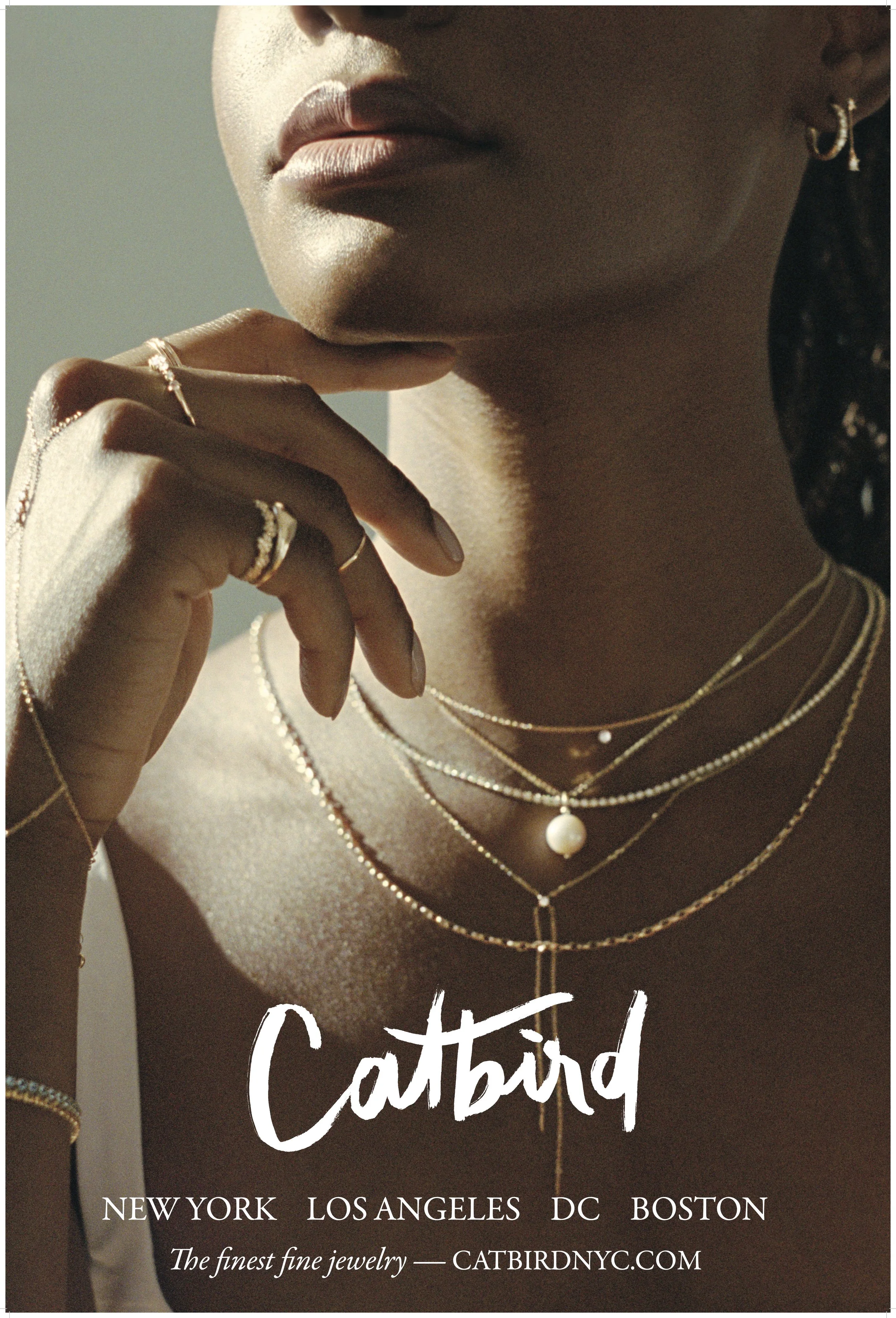 Close-up of a woman wearing layered gold necklaces with pearl pendant, multiple rings, and gold hoop earrings, with her hand near her chin, promoting Catbird jewelry brand in New York, Los Angeles, DC, and Boston.