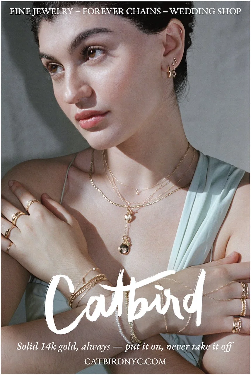 A young woman with brown hair wearing multiple layered gold necklaces, earrings, rings, and bracelets, showcasing jewelry from Catbird, a jewelry store.