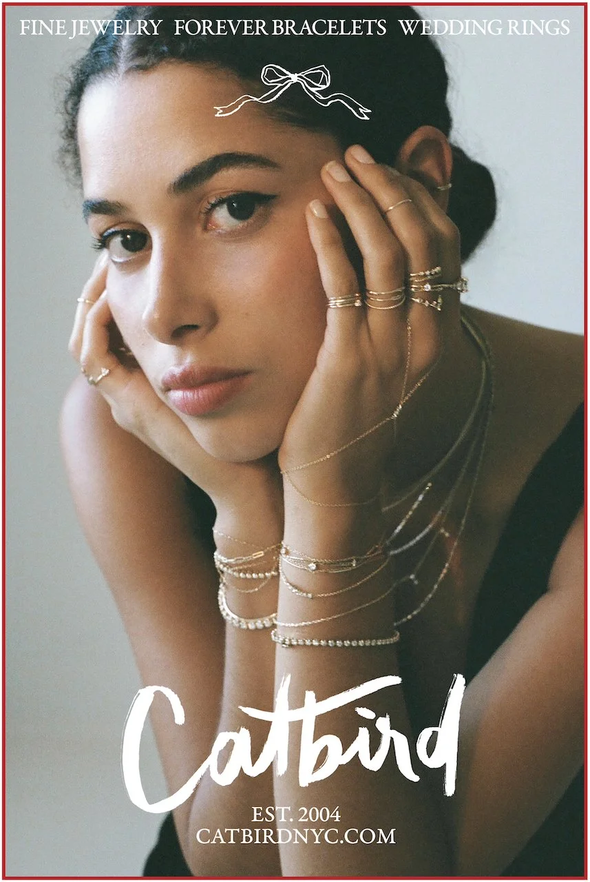 Close-up of a woman with dark curly hair adorned with jewelry, gazing softly at the camera, with her hands resting on her face showing multiple rings and bracelets. The image promotes jewelry, including fine jewelry, bracelets, wedding rings, and Cat