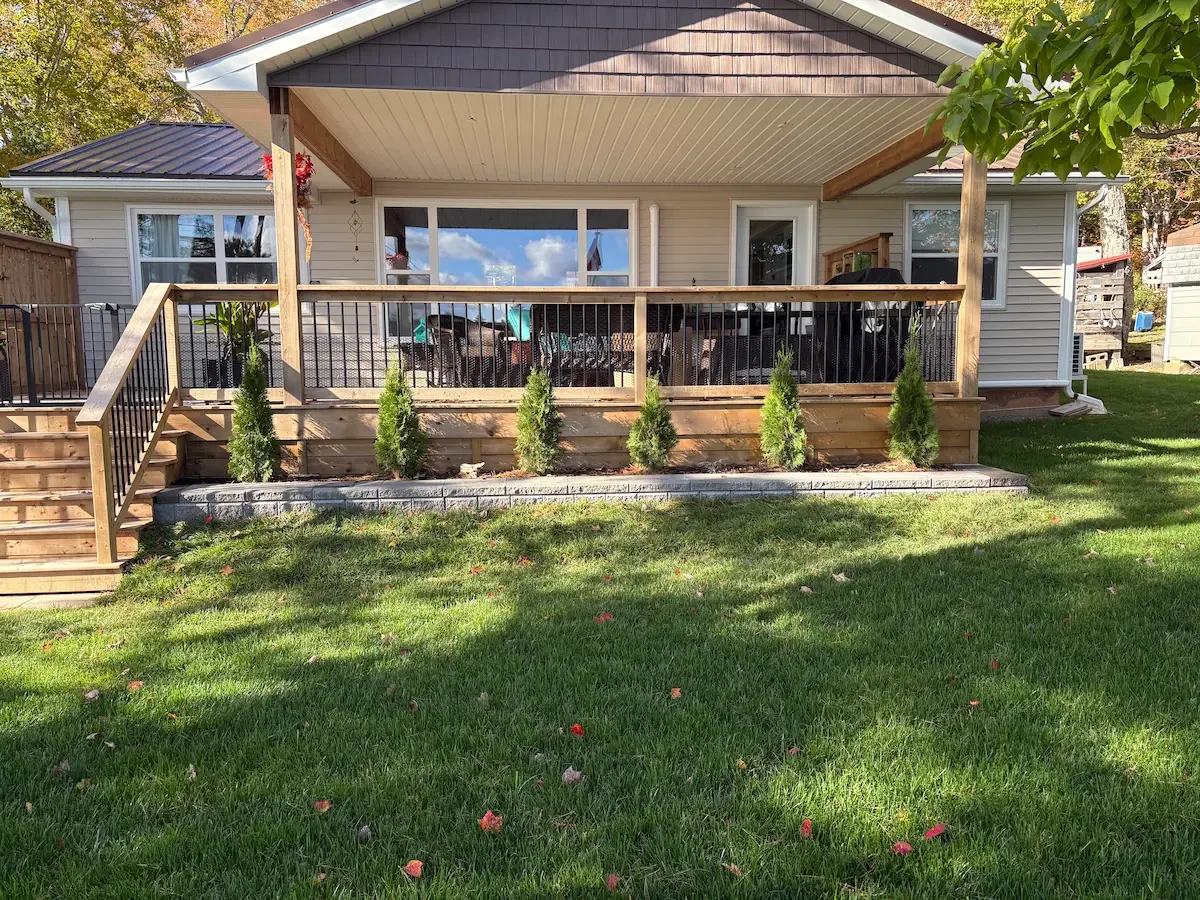 Backyard hardscaping with a raised decorative stone garden bed, decorative evergreen shrubs, and a wooden deck with black railings.