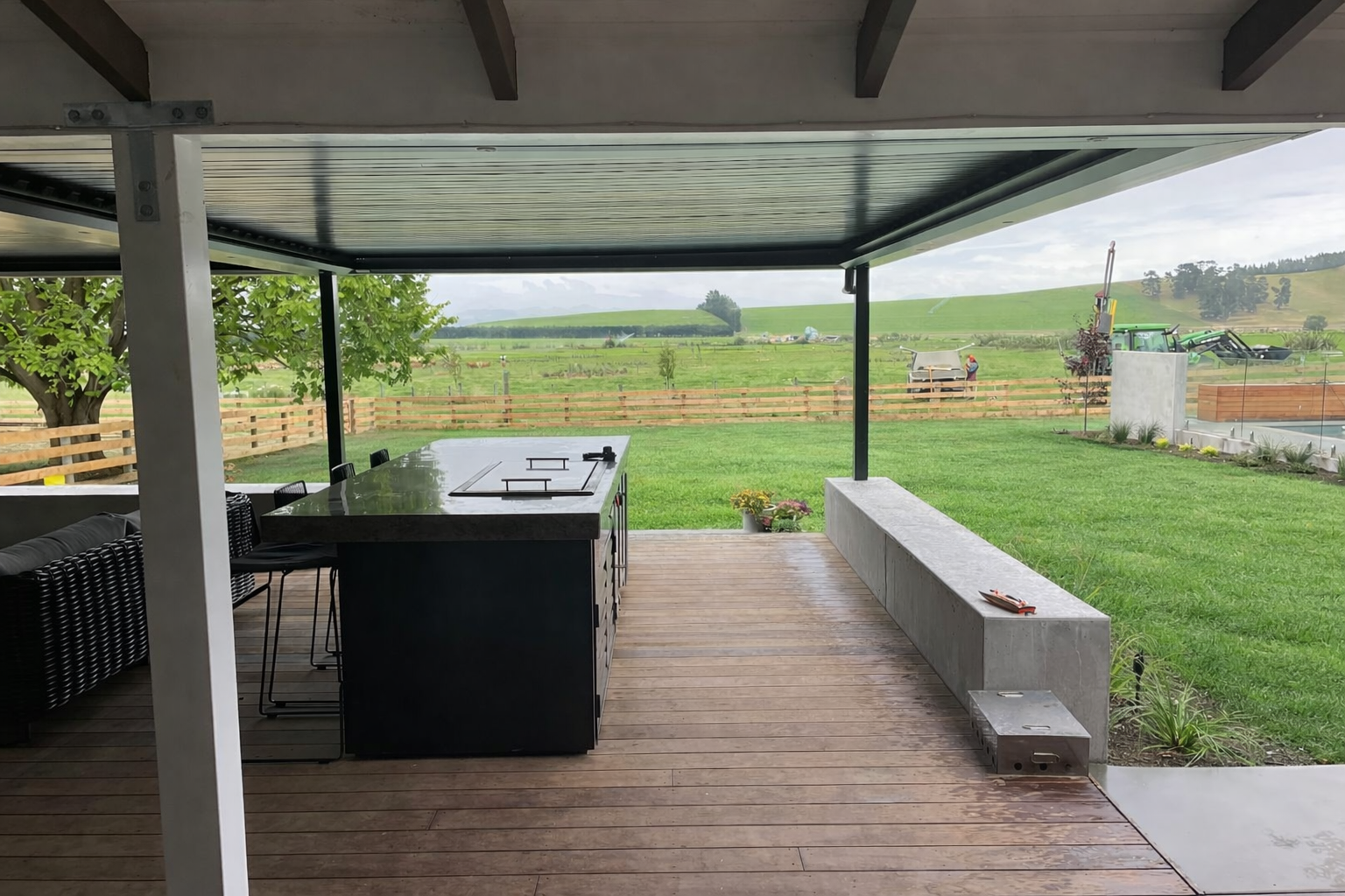 Covered outdoor patio with a high-top table, bar stools, and a view of a green lawn, trees, rolling hills, and a construction site in the distance.