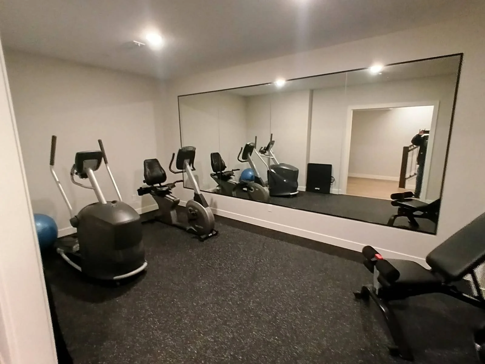 Empty home gym with exercise machines, a large wall mirror, and a person taking a photo in the background.