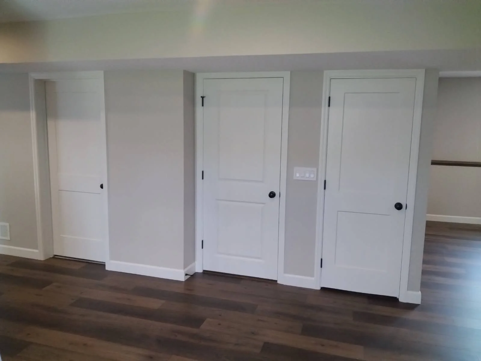 Interior of a room with four closed white doors, light gray walls, brown hardwood flooring, and a white electrical outlet and light switch.