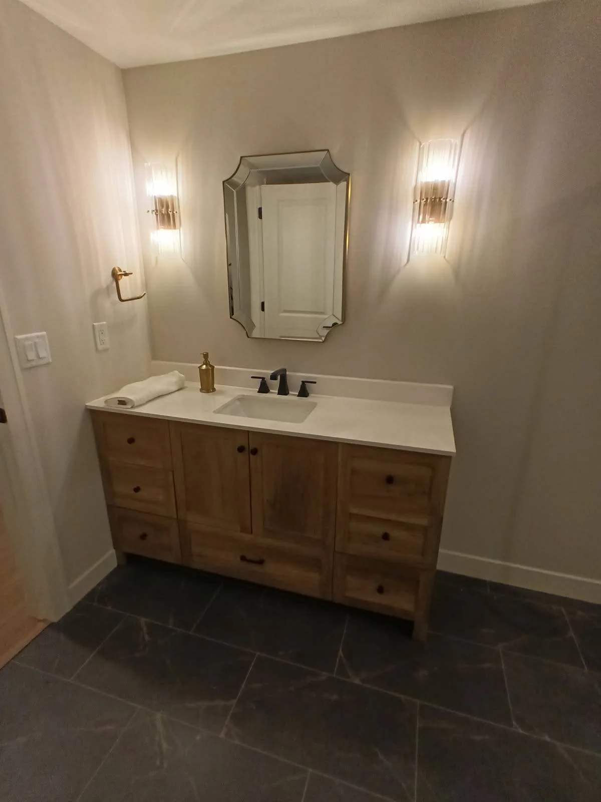 Bathroom vanity with wooden cabinet, white countertop, black faucet, mirror, two wall lights, and a soap dispenser.
