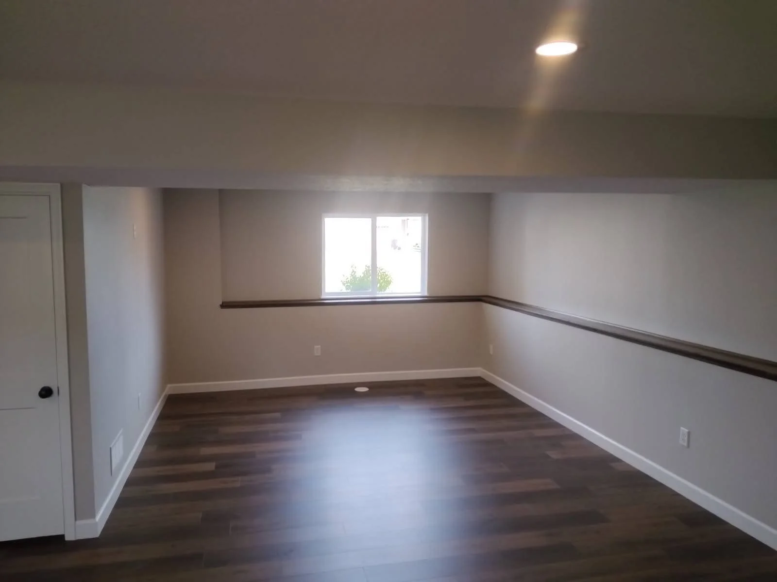 Empty room with wood flooring, white walls, a small window on the back wall, and a ceiling light turned on.