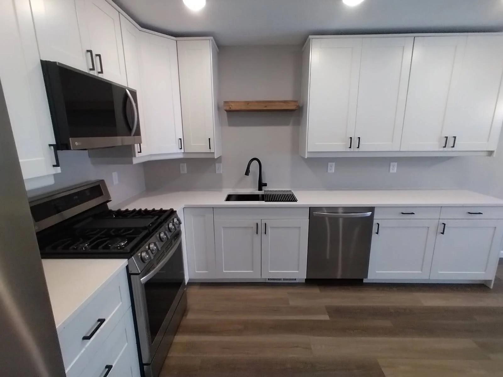 Modern kitchen with white cabinets, stainless steel appliances including a microwave, oven, and dishwasher, black faucet, black handles, and wooden flooring.