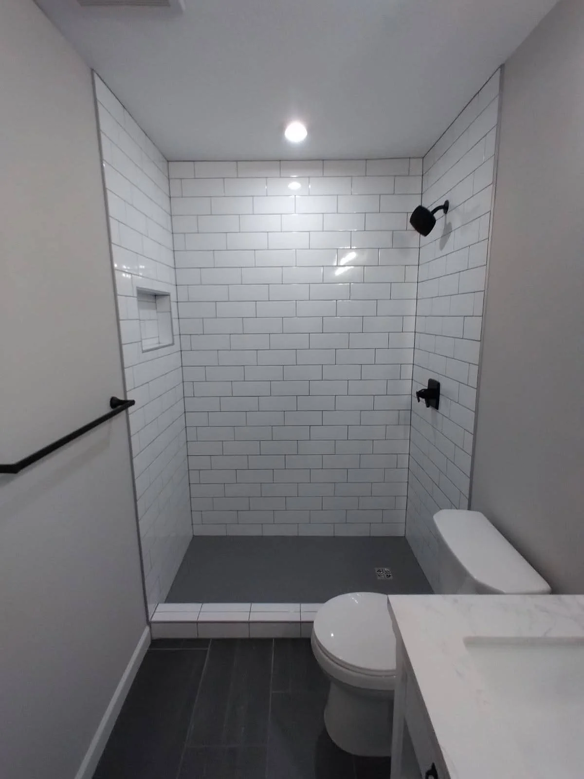Bathroom with walk-in shower featuring white subway tiles, black shower fixtures, a toilet, a marble countertop sink, and a black handrail.