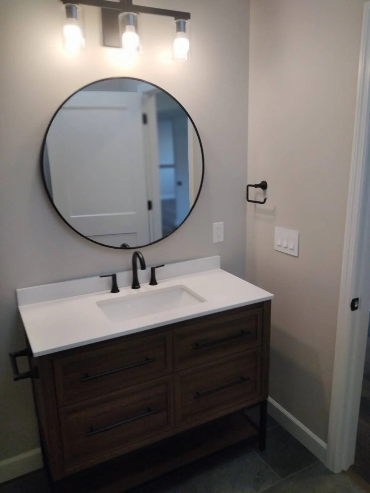 Modern bathroom vanity with a white countertop, black faucet and handles, a round mirror, and four light fixtures above the mirror. The wall is painted a neutral beige color.