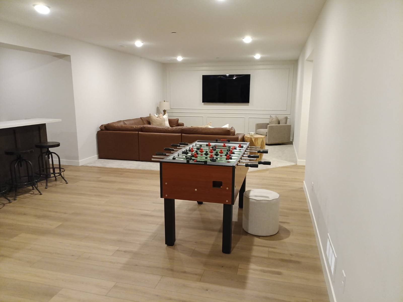 A spacious living room with a foosball table in the center, a brown leather sectional sofa with pillows, a wall-mounted flat-screen TV, and a small beige armchair with a side table in a well-lit area with white walls and hardwood flooring.