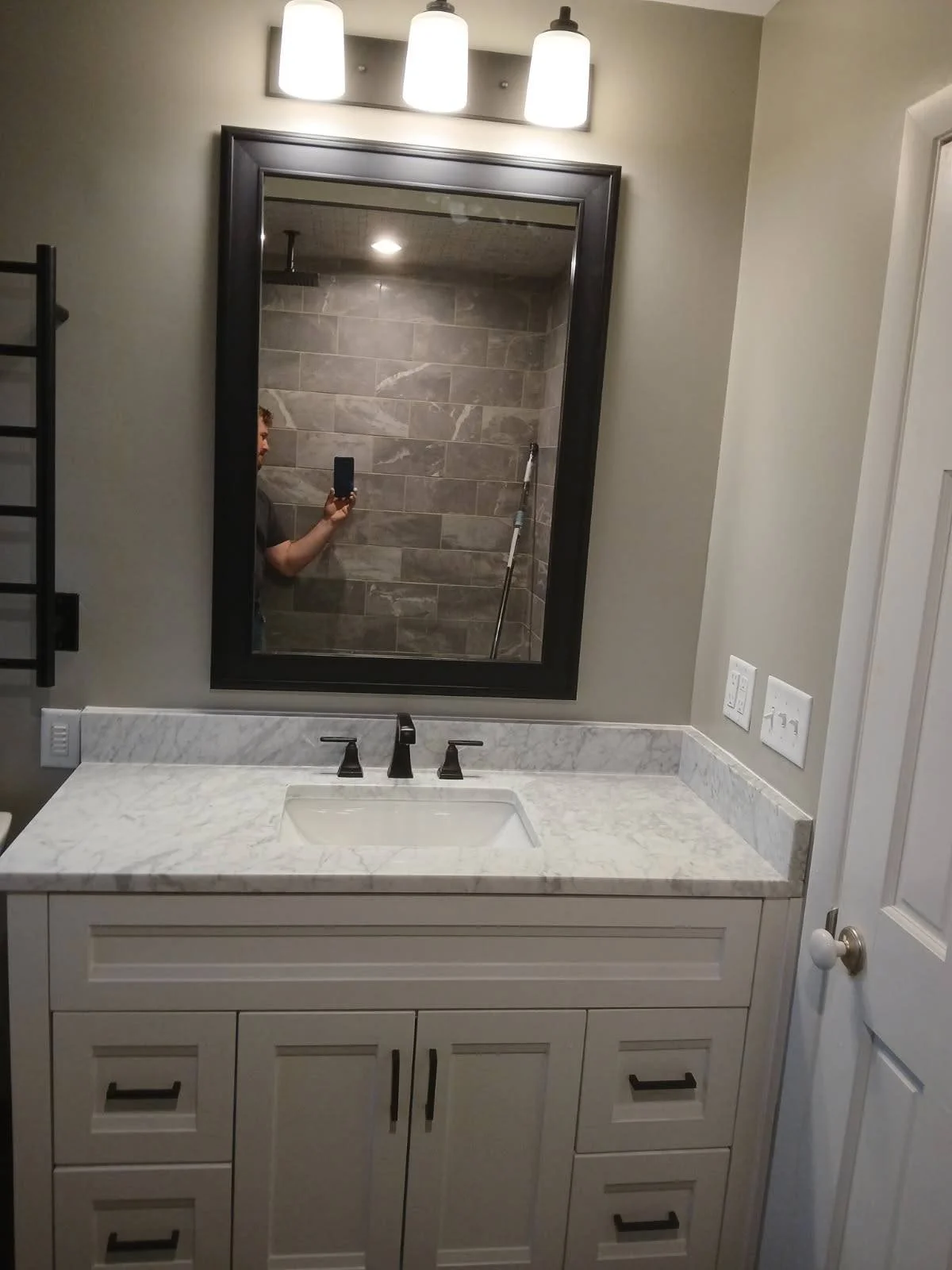 Bathroom vanity with white marble countertop, rectangular sink, black fixtures, large mirror, and wall-mounted light fixture with three bulbs.